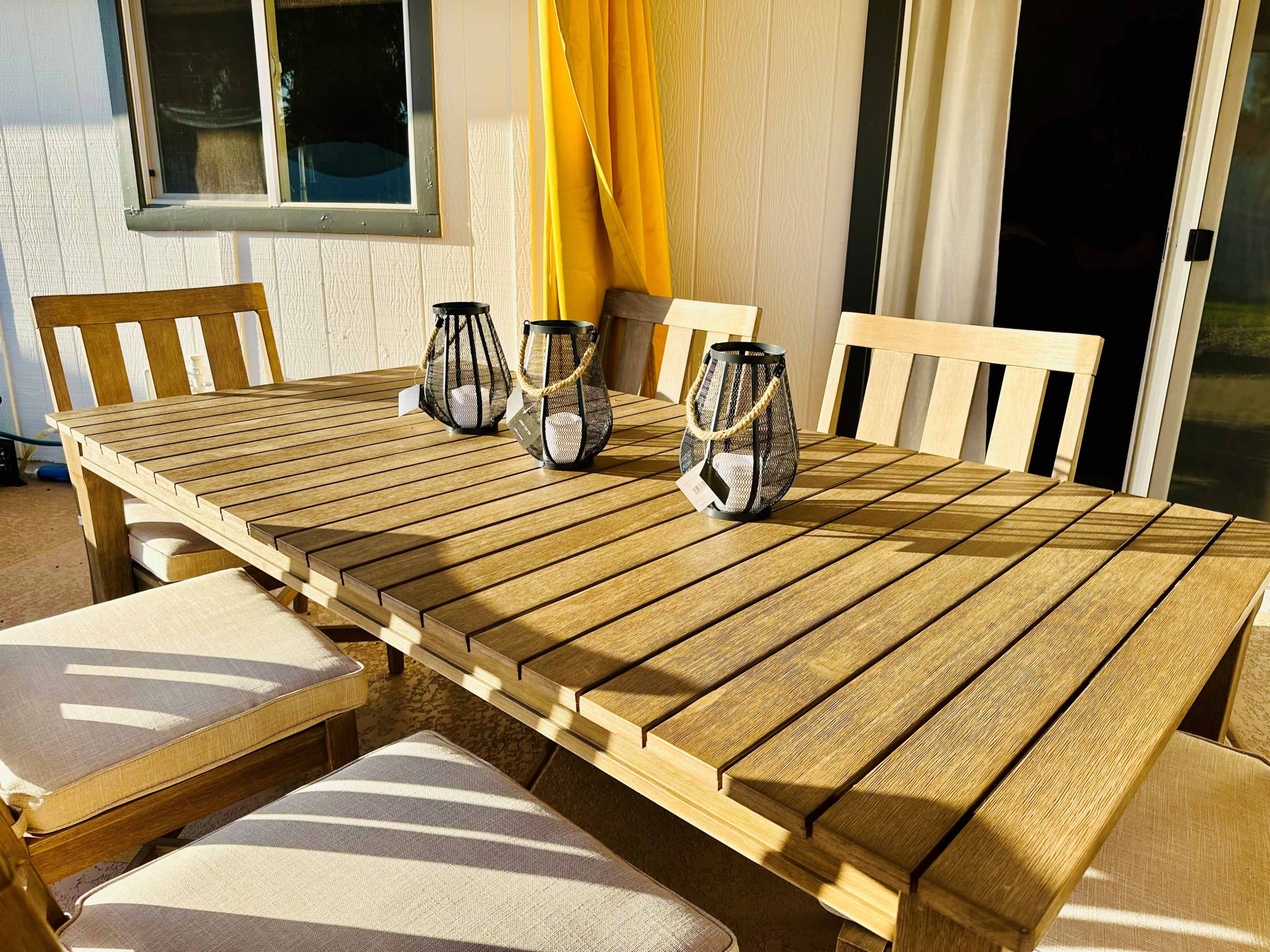 A wooden outdoor dining table is set with three decorative lanterns, accompanied by four cushioned chairs, positioned near a yellow curtain.