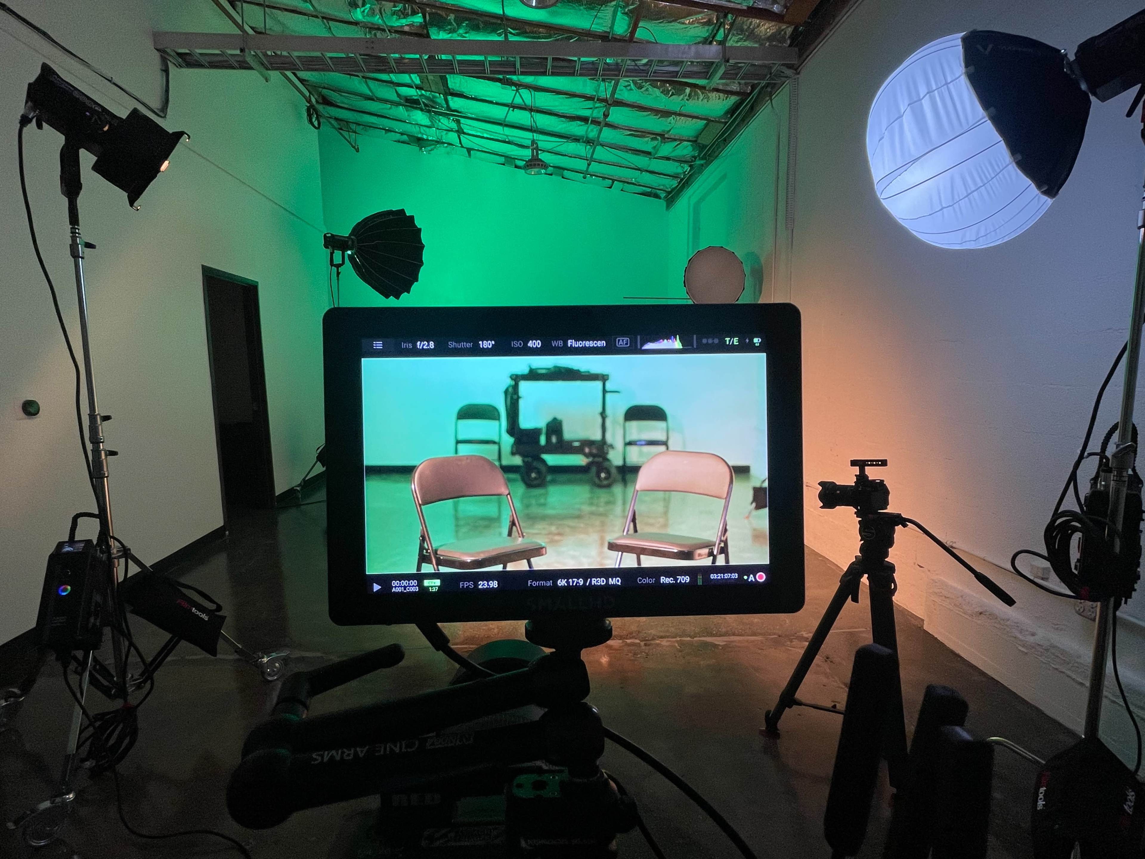 A video monitor displays a scene with empty chairs set up in a studio with various lighting equipment.