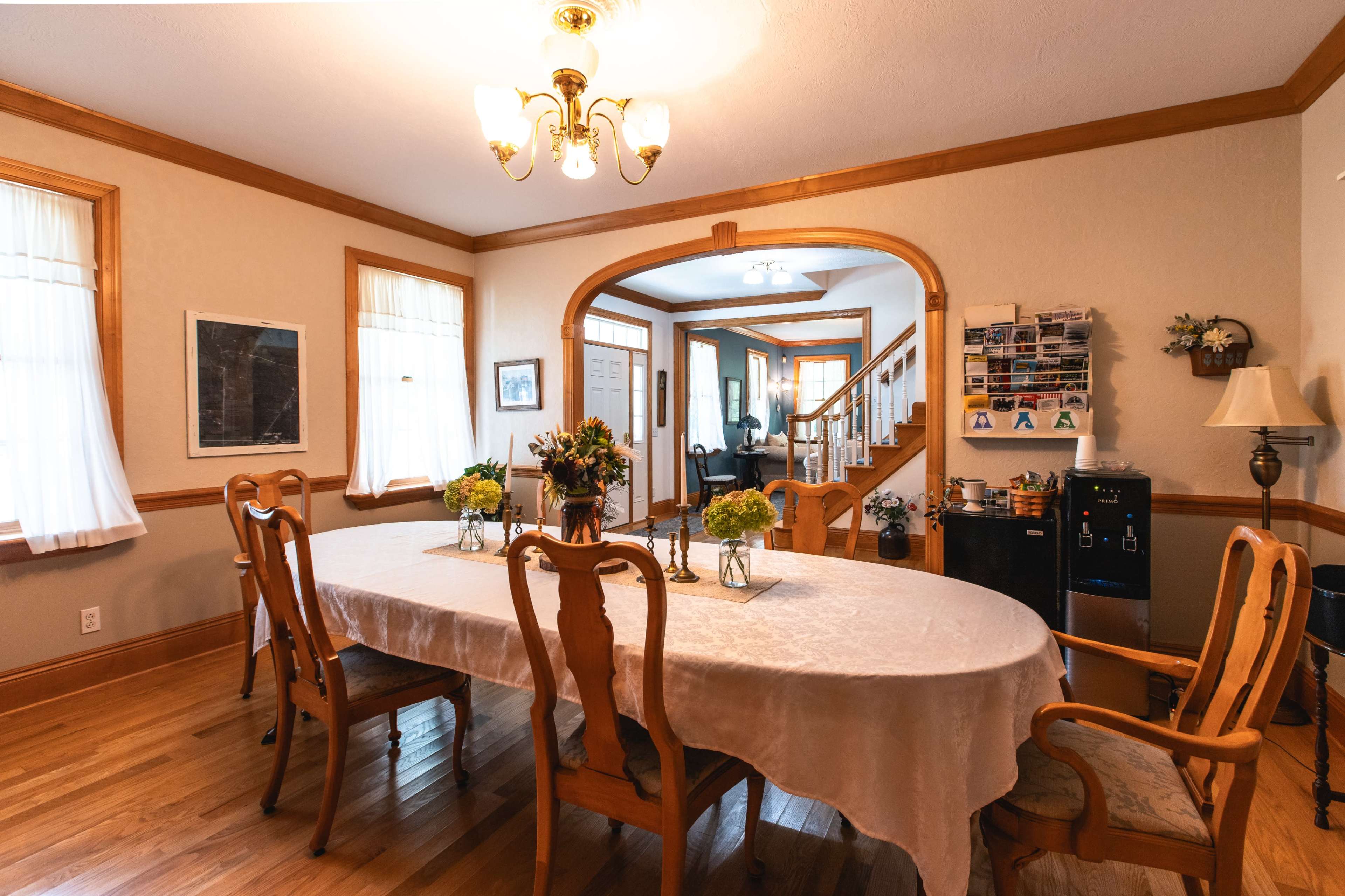 The image shows a dining room with a long table surrounded by wooden chairs, featuring a chandelier overhead and large windows allowing natural light.
