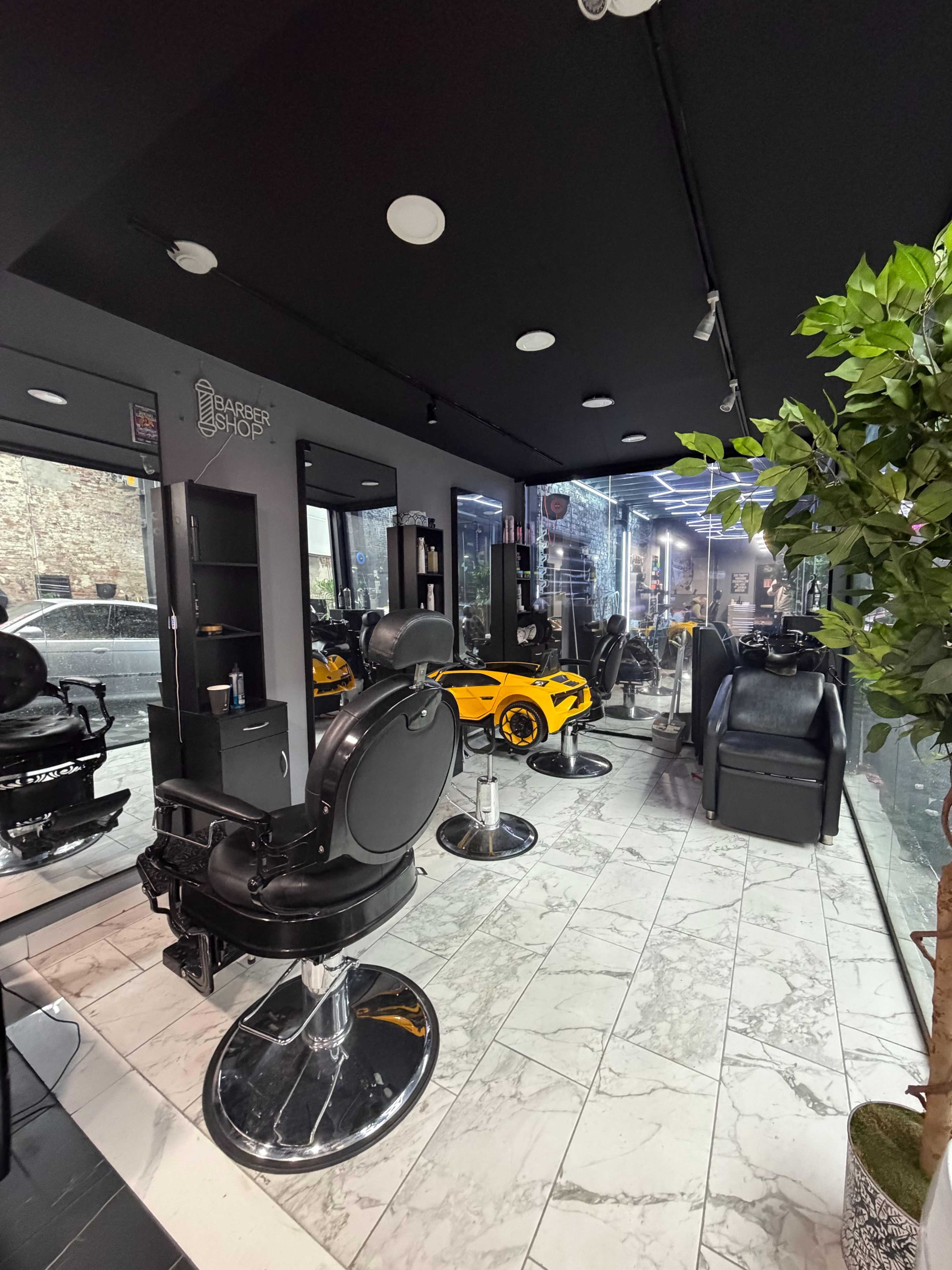 The image shows a modern barbershop interior featuring several black barber chairs, mirrors, and a yellow sports car placed in the corner.