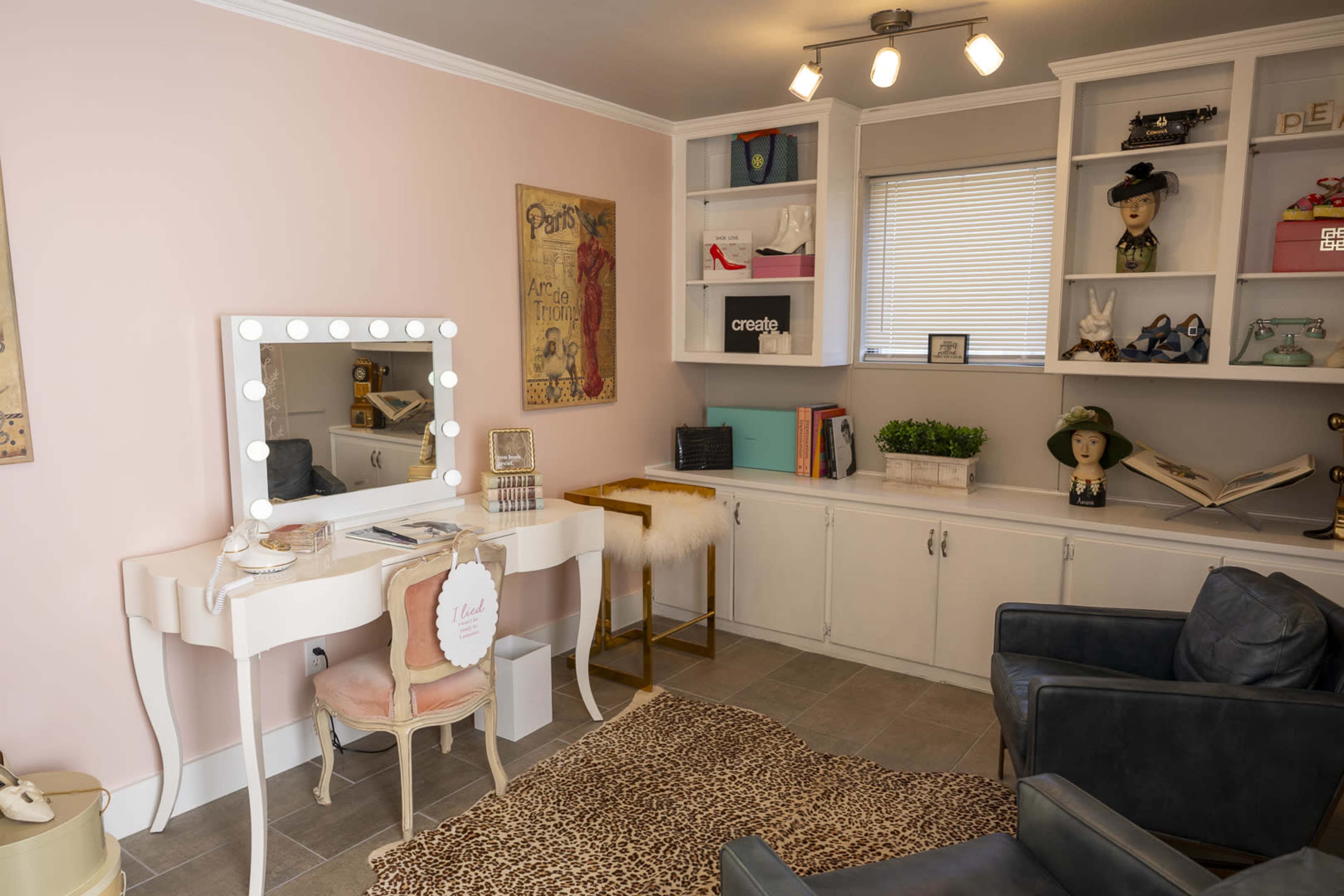 The image shows a stylish home office with a vanity desk, a black armchair, shelves with decorative items, and a light pink wall.