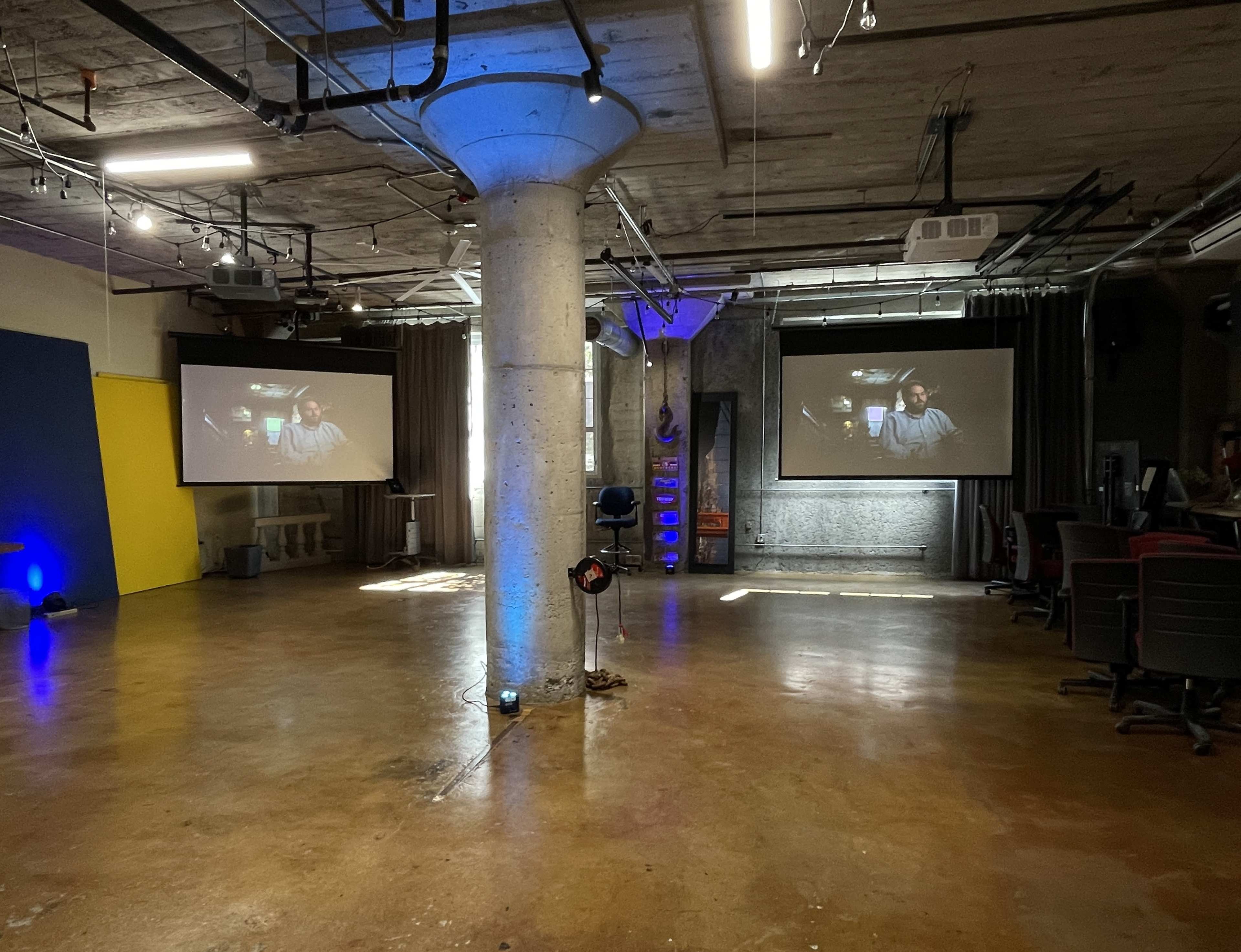 The image shows a spacious room with two projection screens displaying video, a concrete pillar in the center, and various seating arrangements along the sides.
