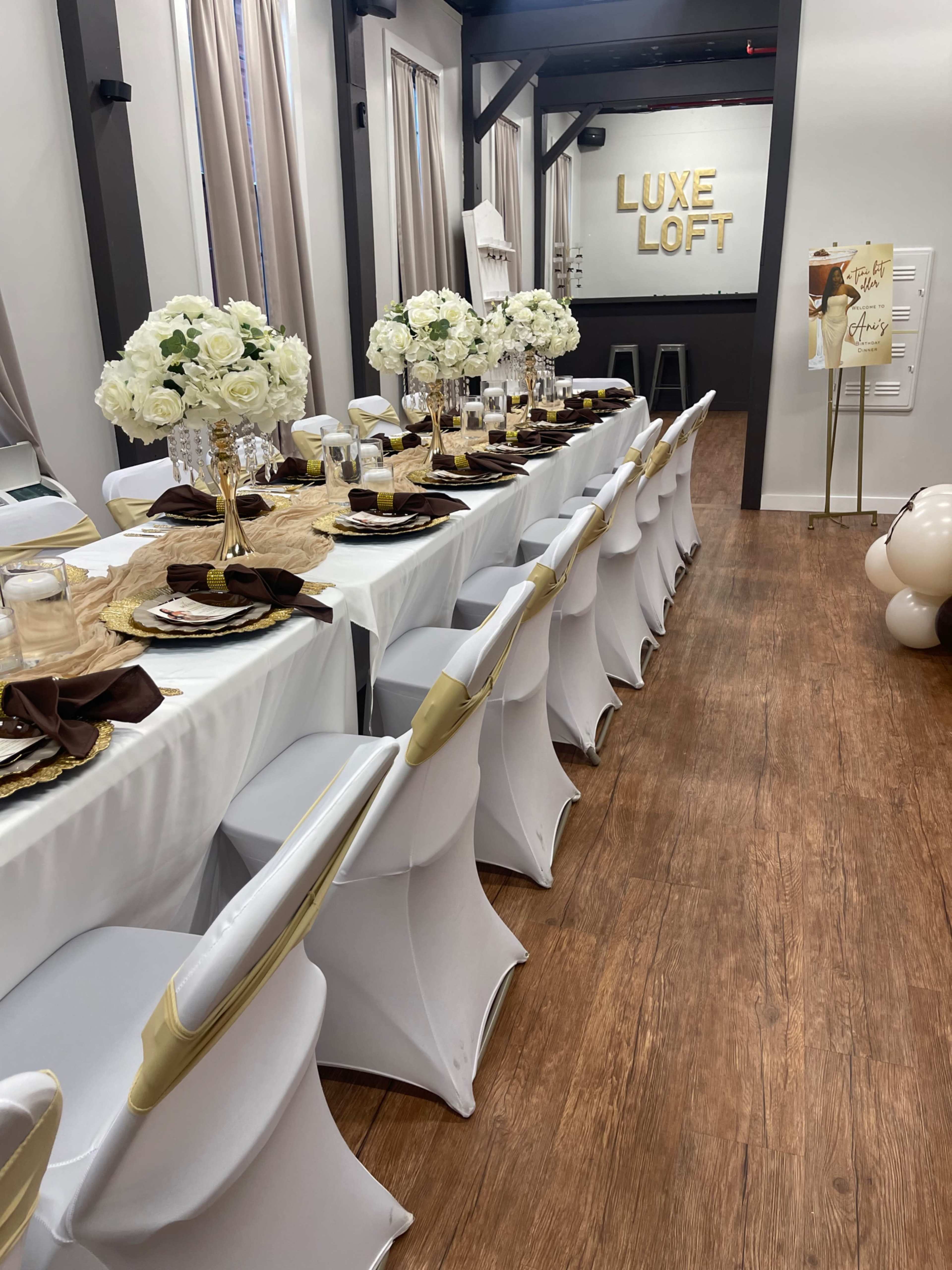 A long dining table is set with white tablecloths, elegant floral centerpieces, and gold accents in a venue featuring the words "LUXE LOFT" on the wall.