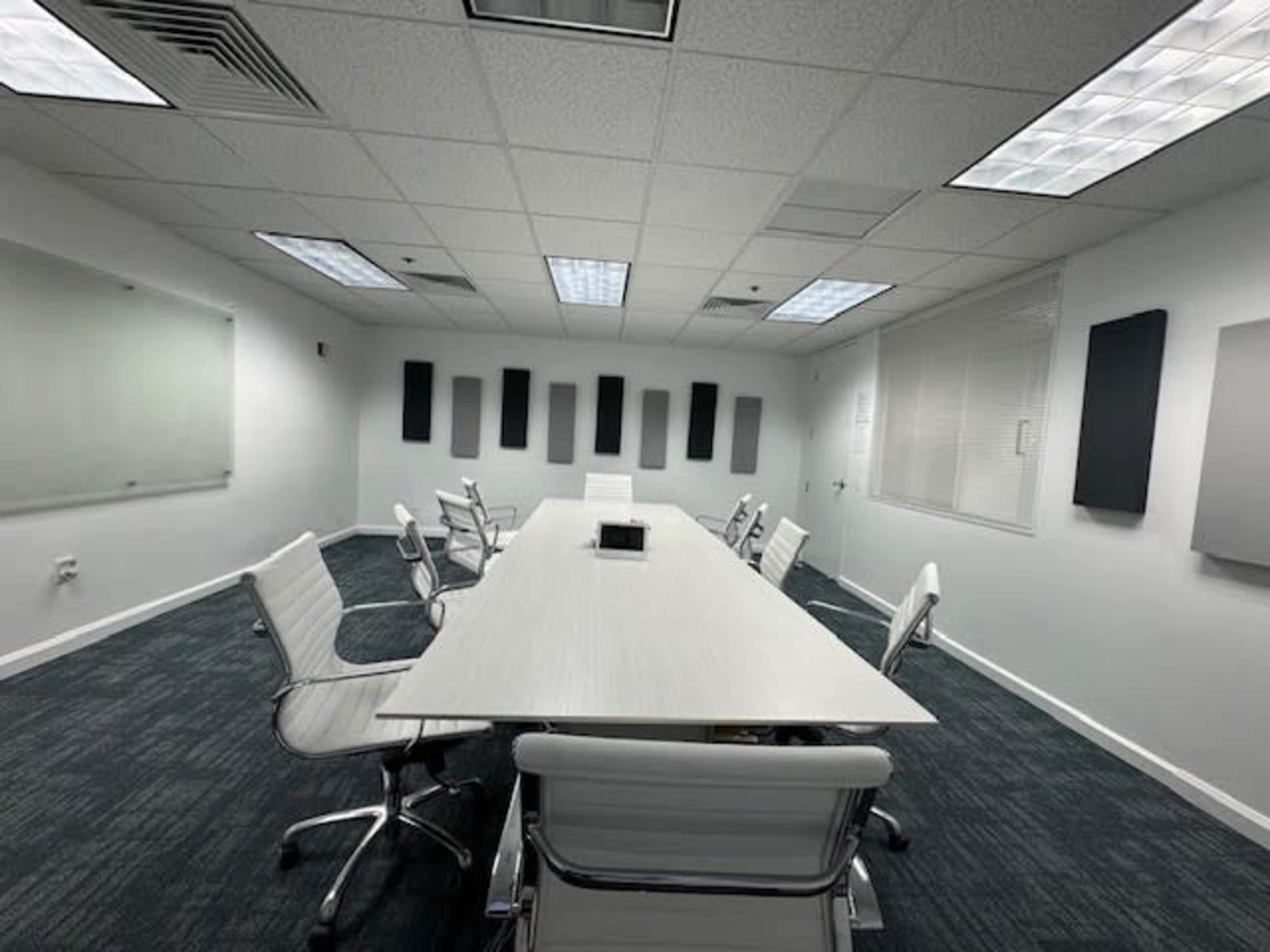 A conference room features a long white table surrounded by sleek chairs, with soundproof panels on the walls and large windows for natural light.