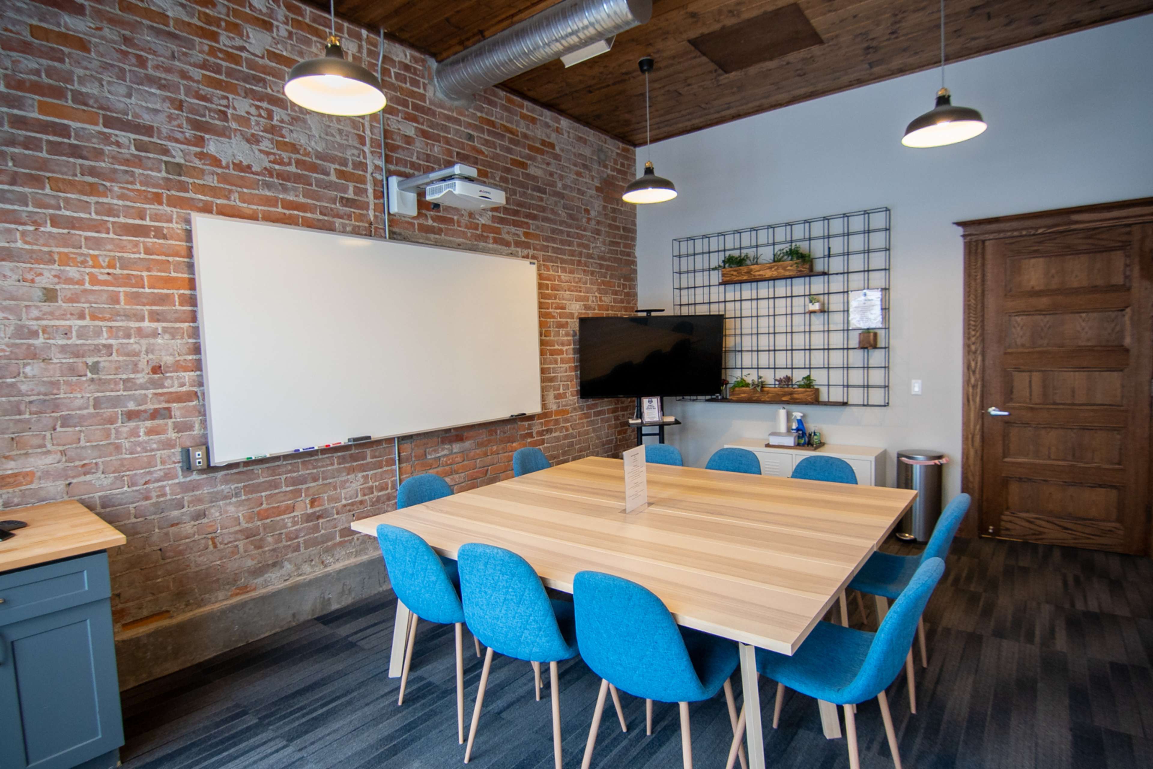 A modern conference room features a wooden table surrounded by blue chairs, a whiteboard, a TV, and exposed brick walls.