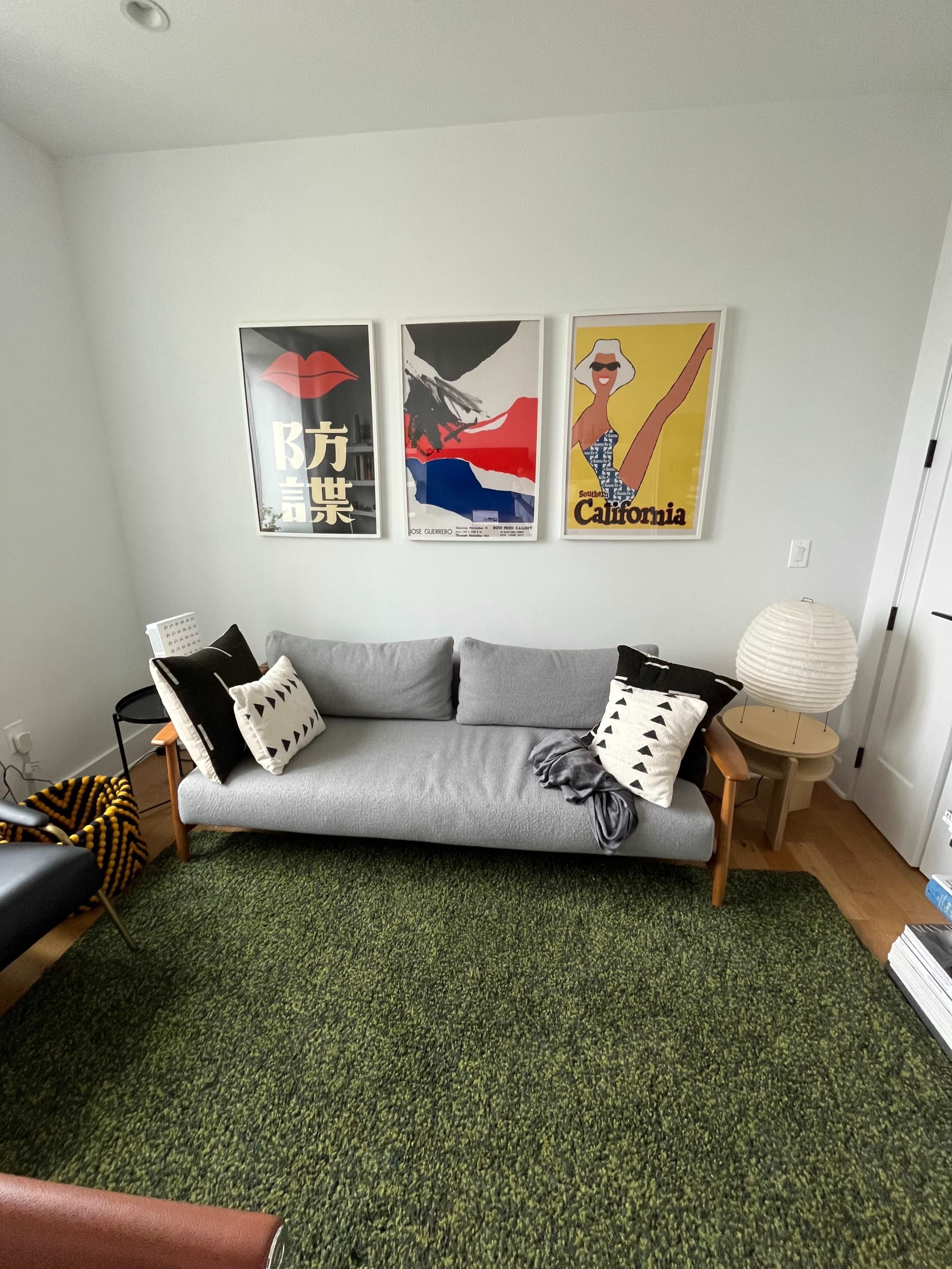 A minimalist living area with a gray sofa, three framed posters on the wall, a round lamp, and a green area rug.