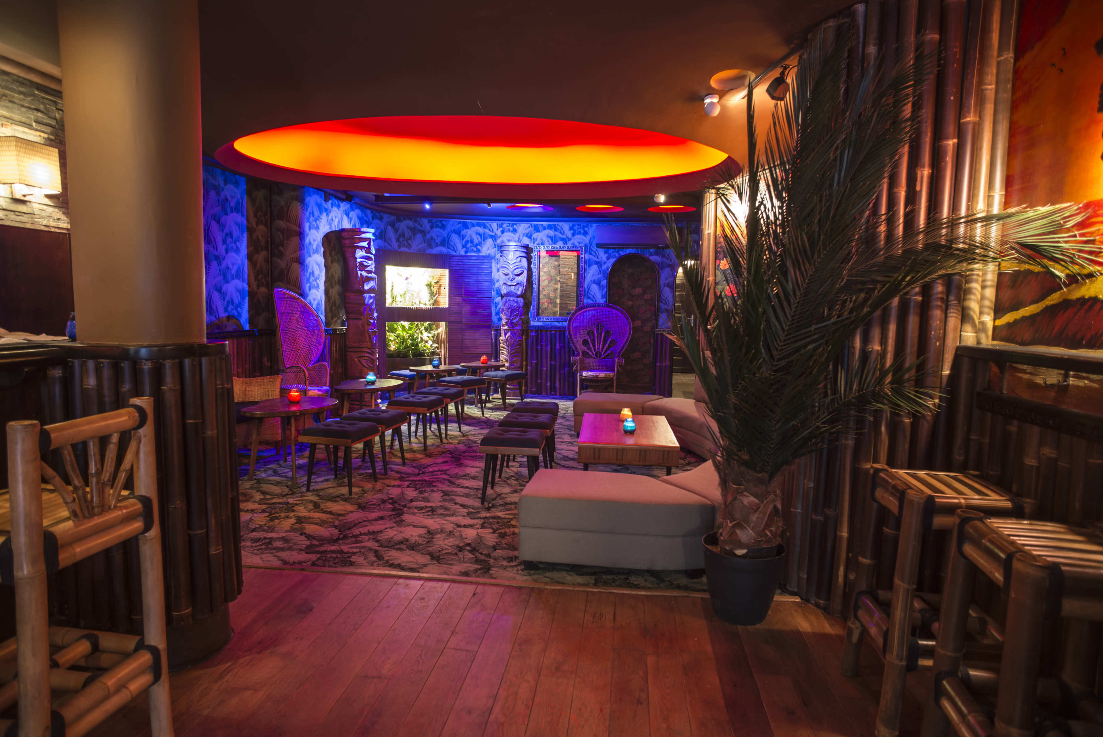 The image shows a dimly lit lounge with bamboo furniture, colorful lighting, and a tropical plant, creating an inviting atmosphere.