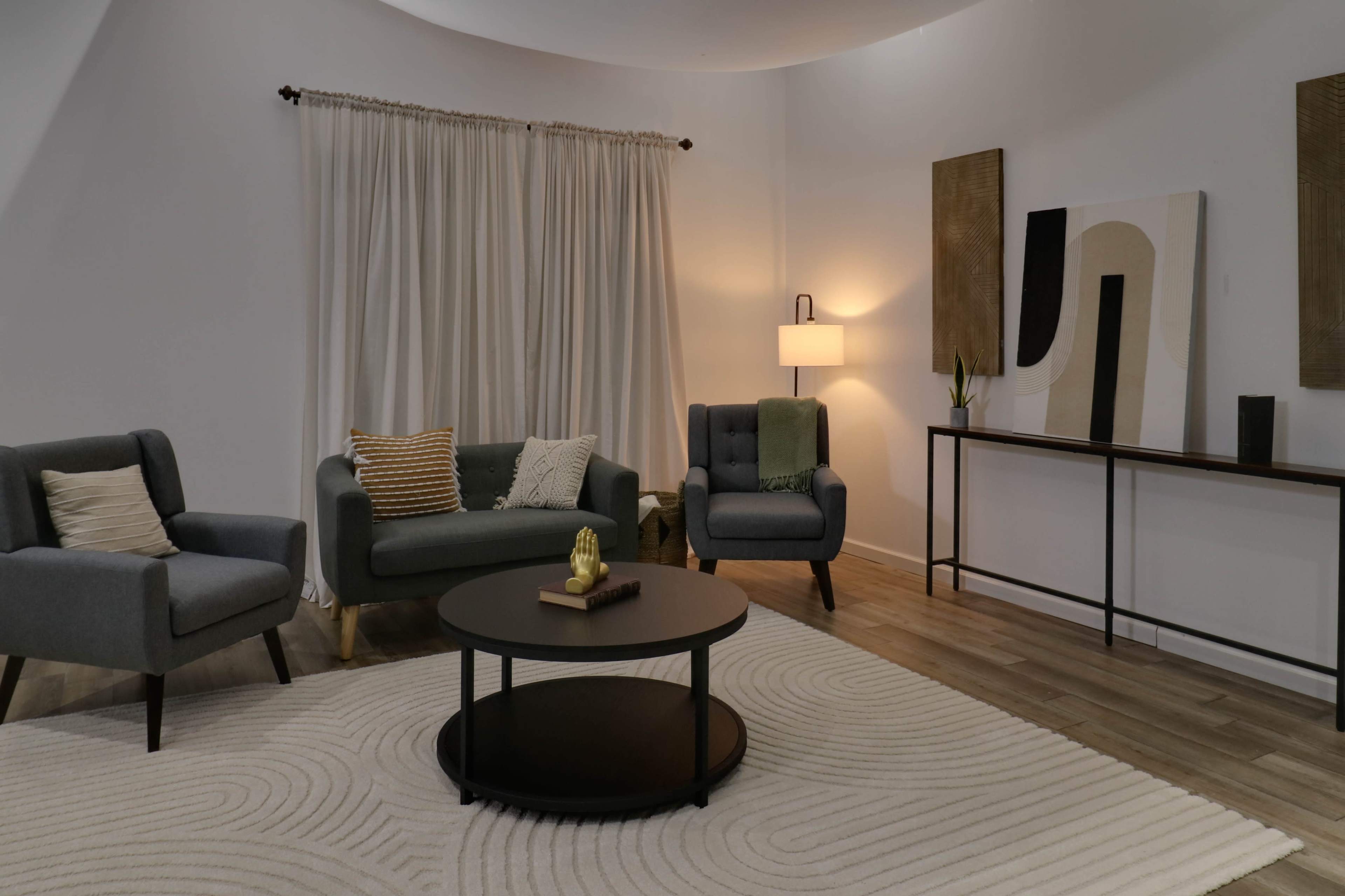 The image shows a modern living room with a gray sofa, two armchairs, a round coffee table, and a minimalist decor featuring a light rug and abstract wall art.
