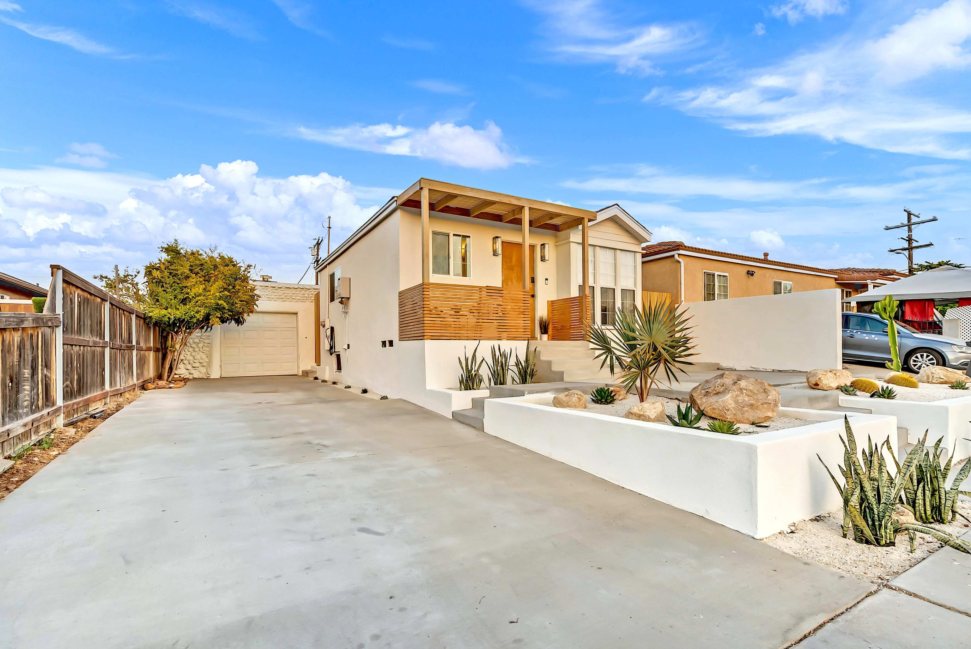 A modern single-story house with a wooden front porch is situated on a concrete driveway surrounded by landscaping featuring rocks and succulents.