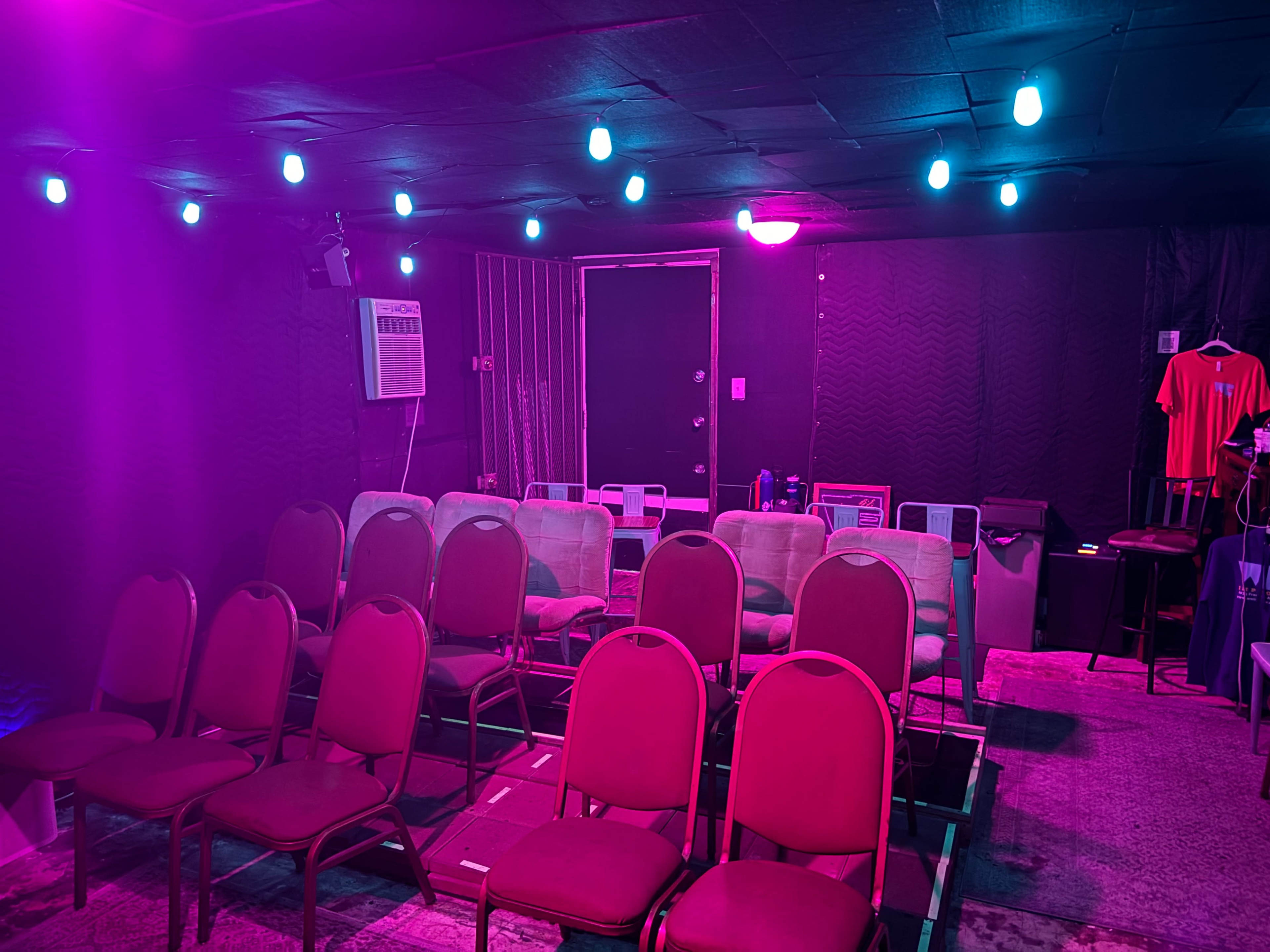 The scene shows a small, dimly lit room set up with rows of empty red chairs facing a door, illuminated by colorful overhead lights.