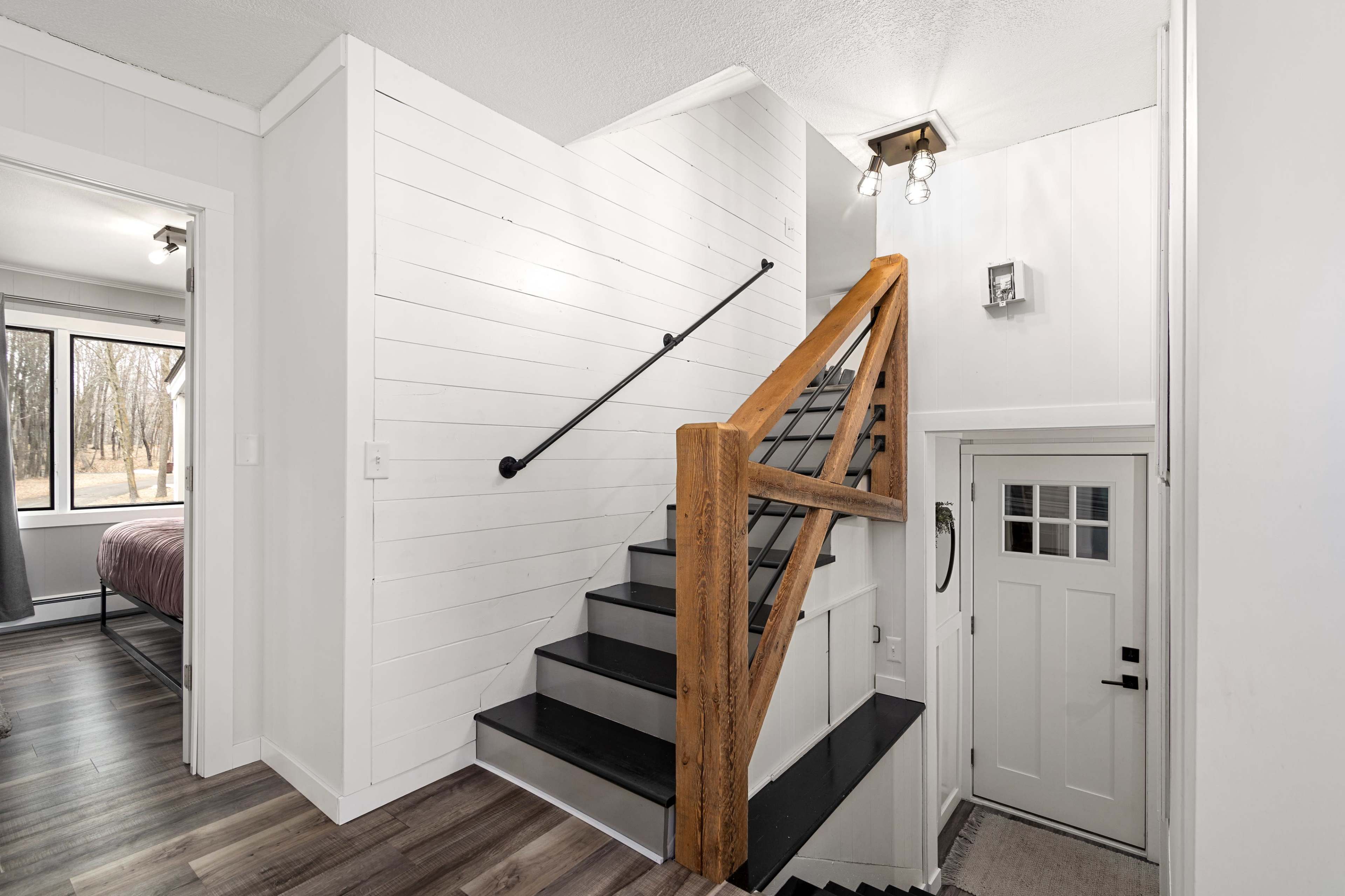 A staircase with wooden handrails leading to a second floor in a modern home, with a front door adjacent to it and large windows in the background.