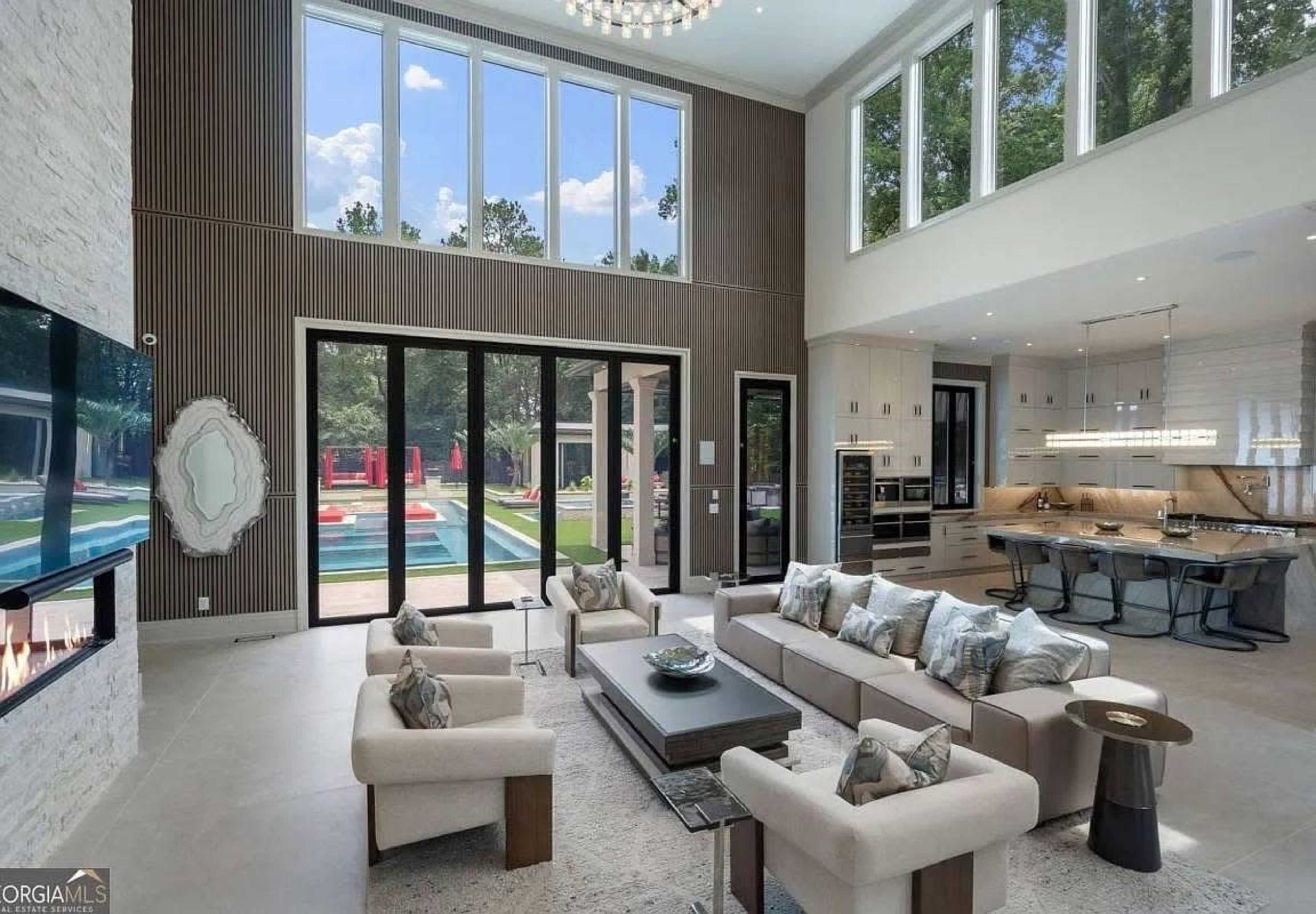 The image shows a spacious, modern living room featuring large windows that provide a view of an outdoor pool area.