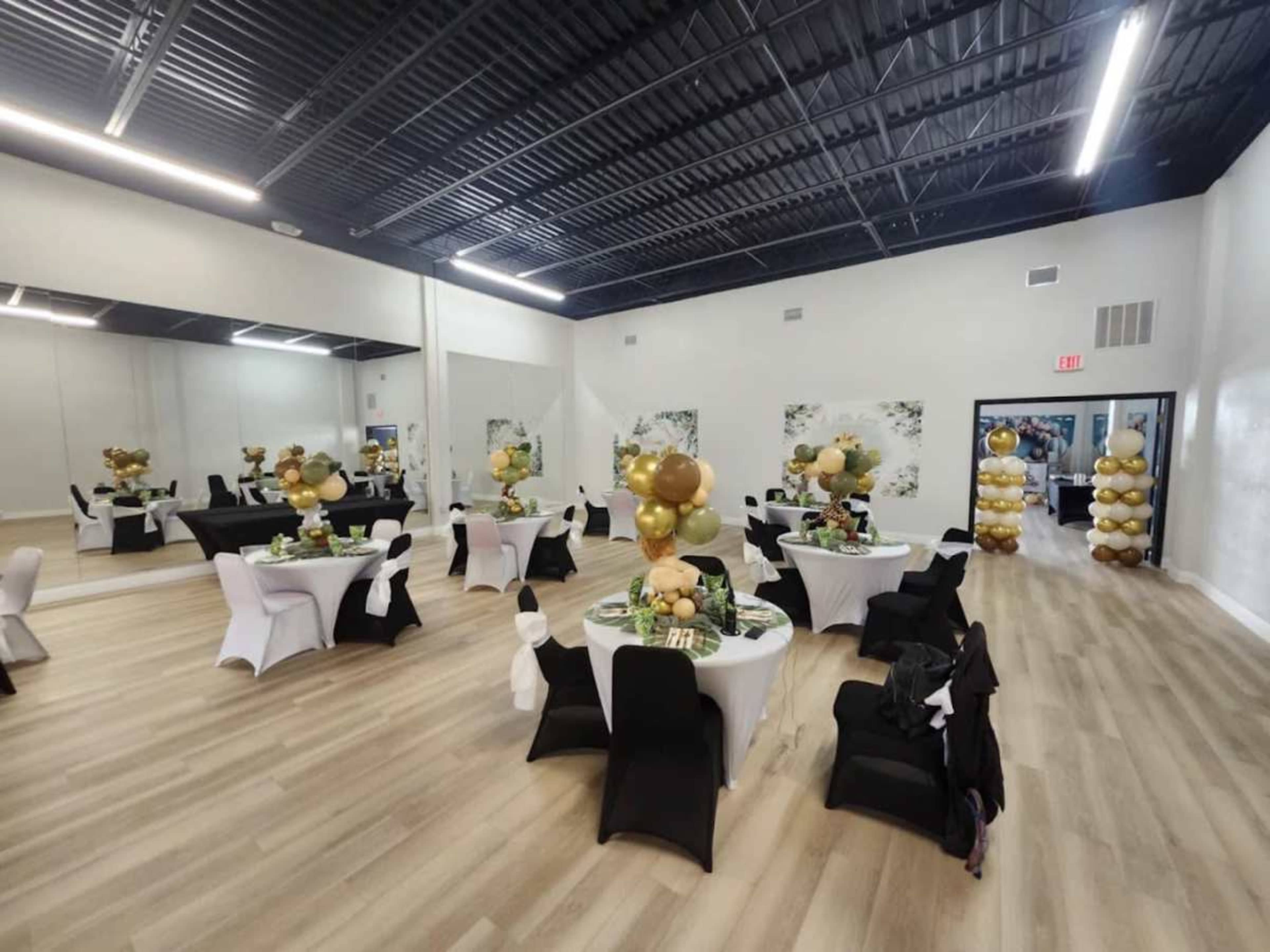 A banquet hall with round tables dressed in white and black tablecloths, decorated with balloons and centerpieces, and a wall of balloons in gold and white at one corner.