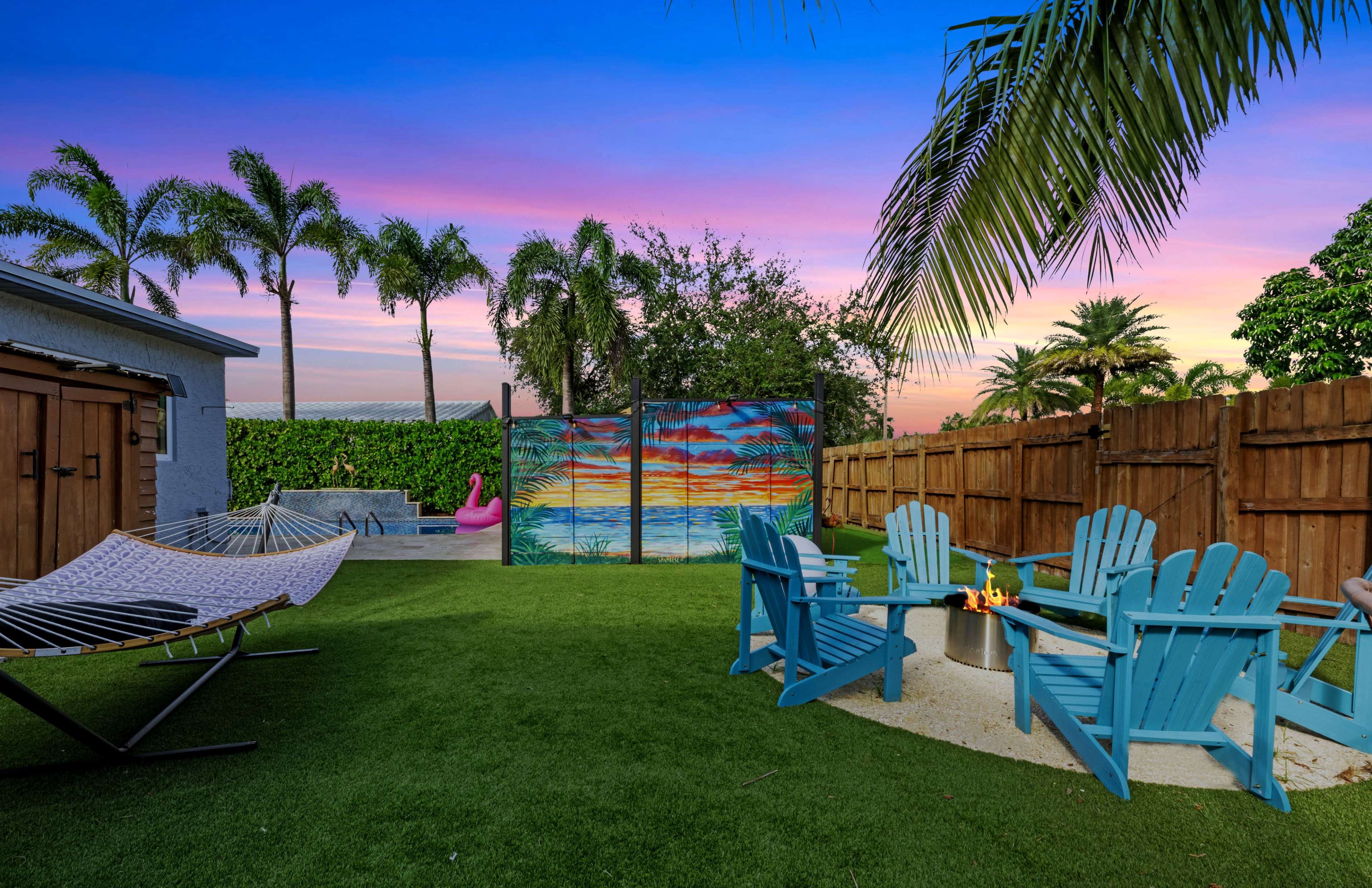 A backyard scene features a hammock, Adirondack chairs surrounding a fire pit, and a colorful mural against a backdrop of palm trees and a sunset sky.