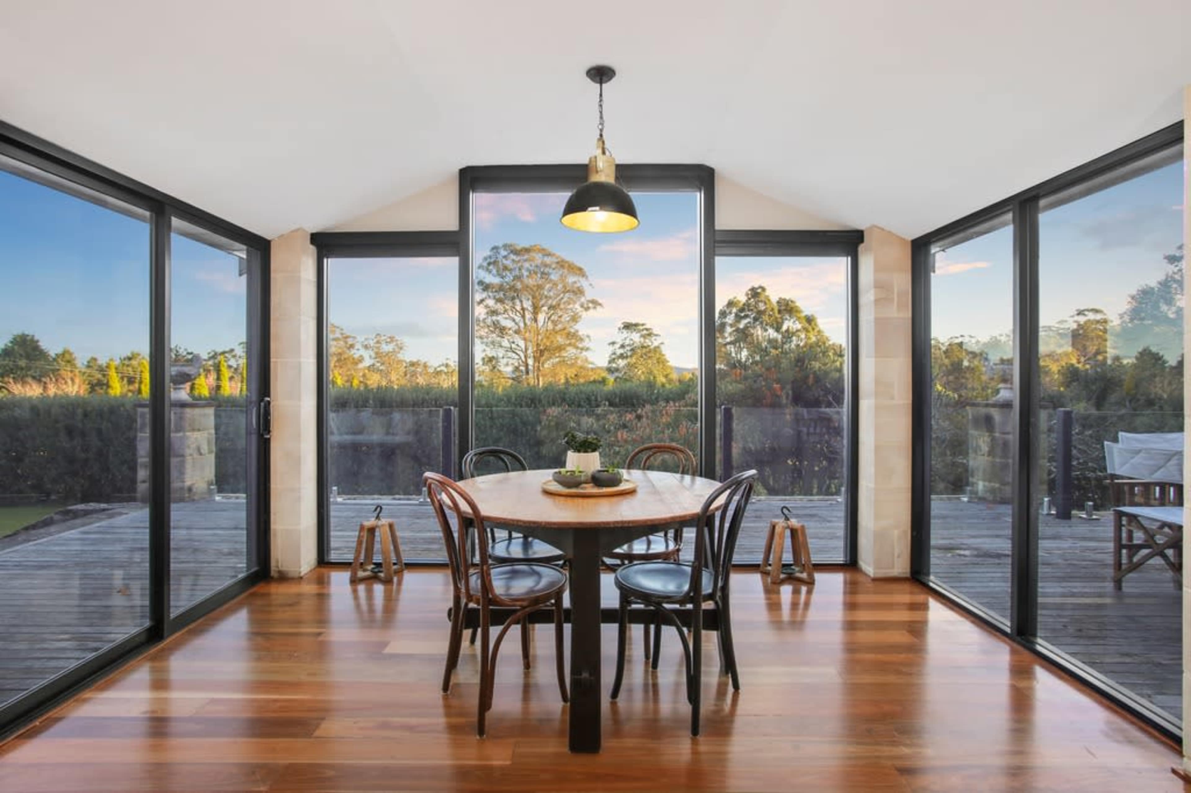 A dining area with a round table and chairs features large glass windows that provide a view of the landscape outside.