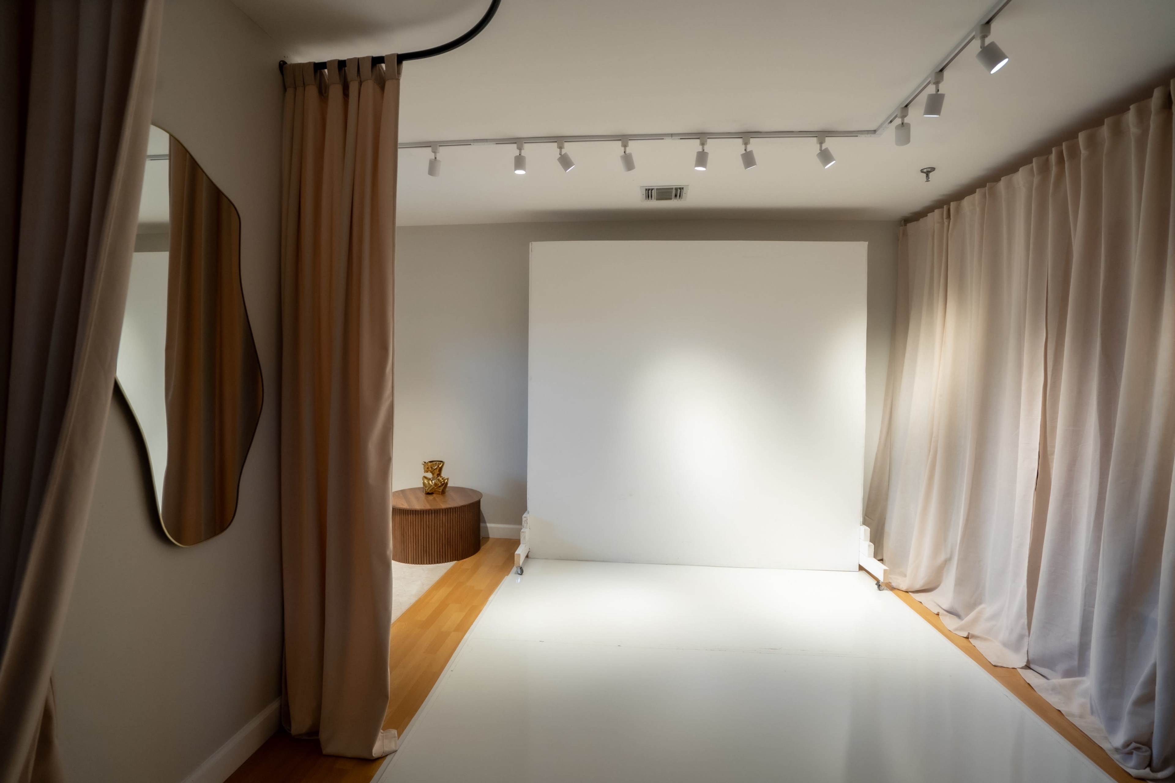 The image shows a minimalist photography studio with a large white backdrop, soft beige curtains, and a round wooden table featuring a small decorative item.