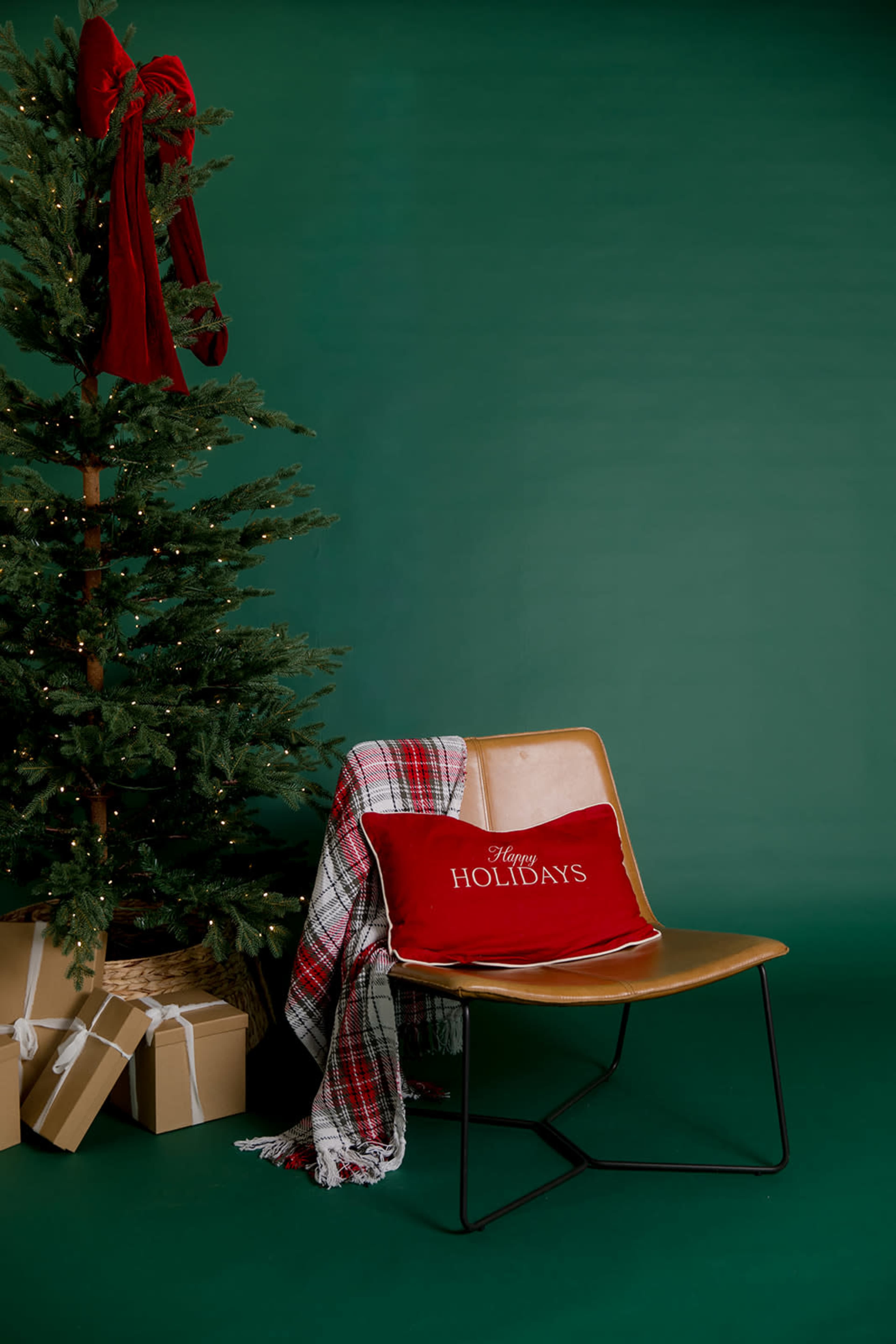 A green background features a Christmas tree adorned with lights and a red ribbon, while a wooden chair holds a red "Happy Holidays" pillow and a plaid blanket, alongside wrapped presents.