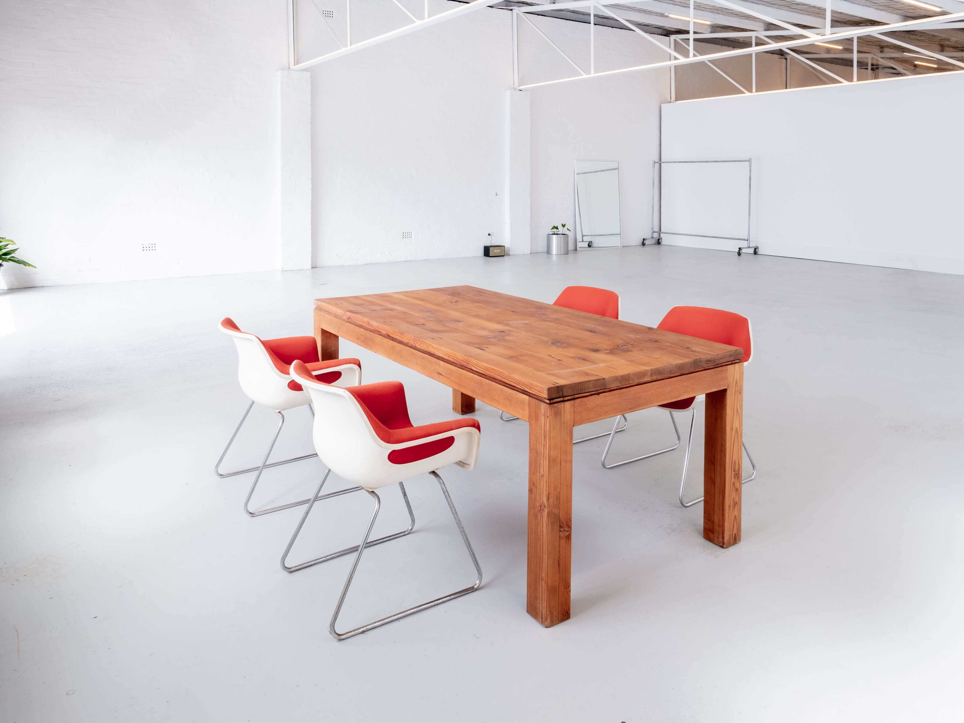 A wooden table with four chairs, two white and two red, is set in a spacious, empty room with white walls and a polished floor.