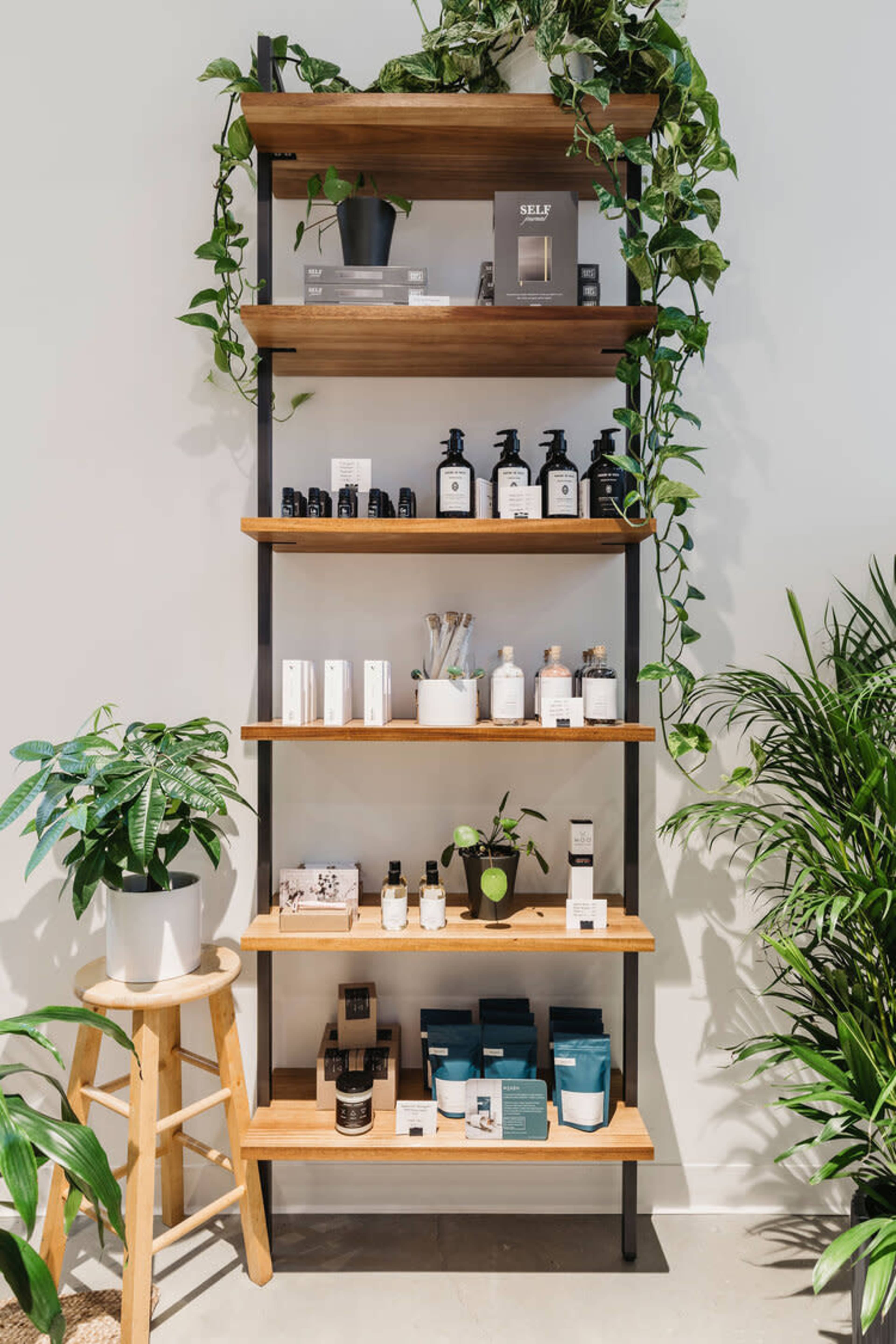 A wooden shelving unit with five shelves displays various skincare products and plants against a white wall.