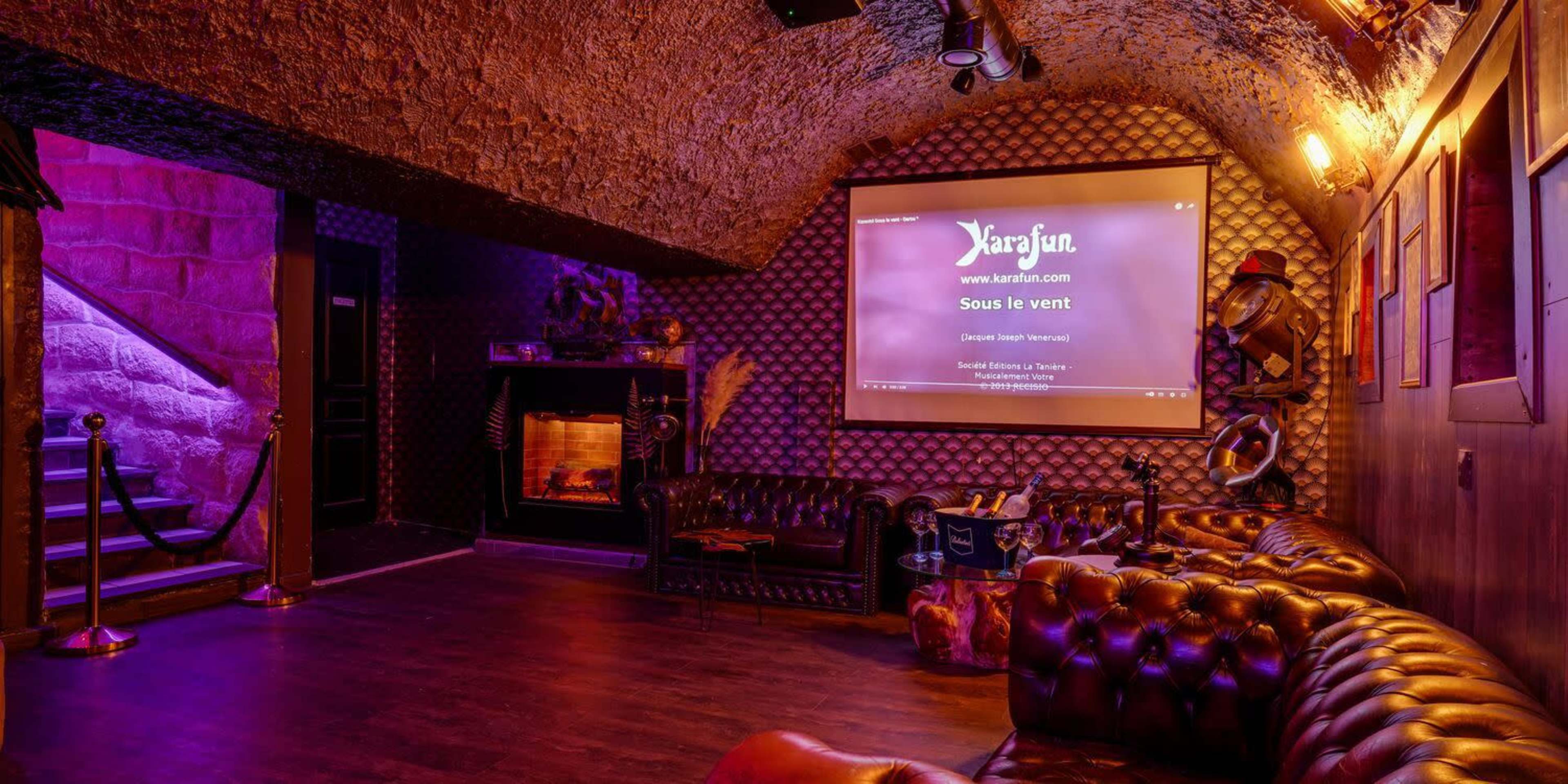 The image shows a dimly lit karaoke lounge with a large screen displaying lyrics, leather seating, and a staircase leading to another area.