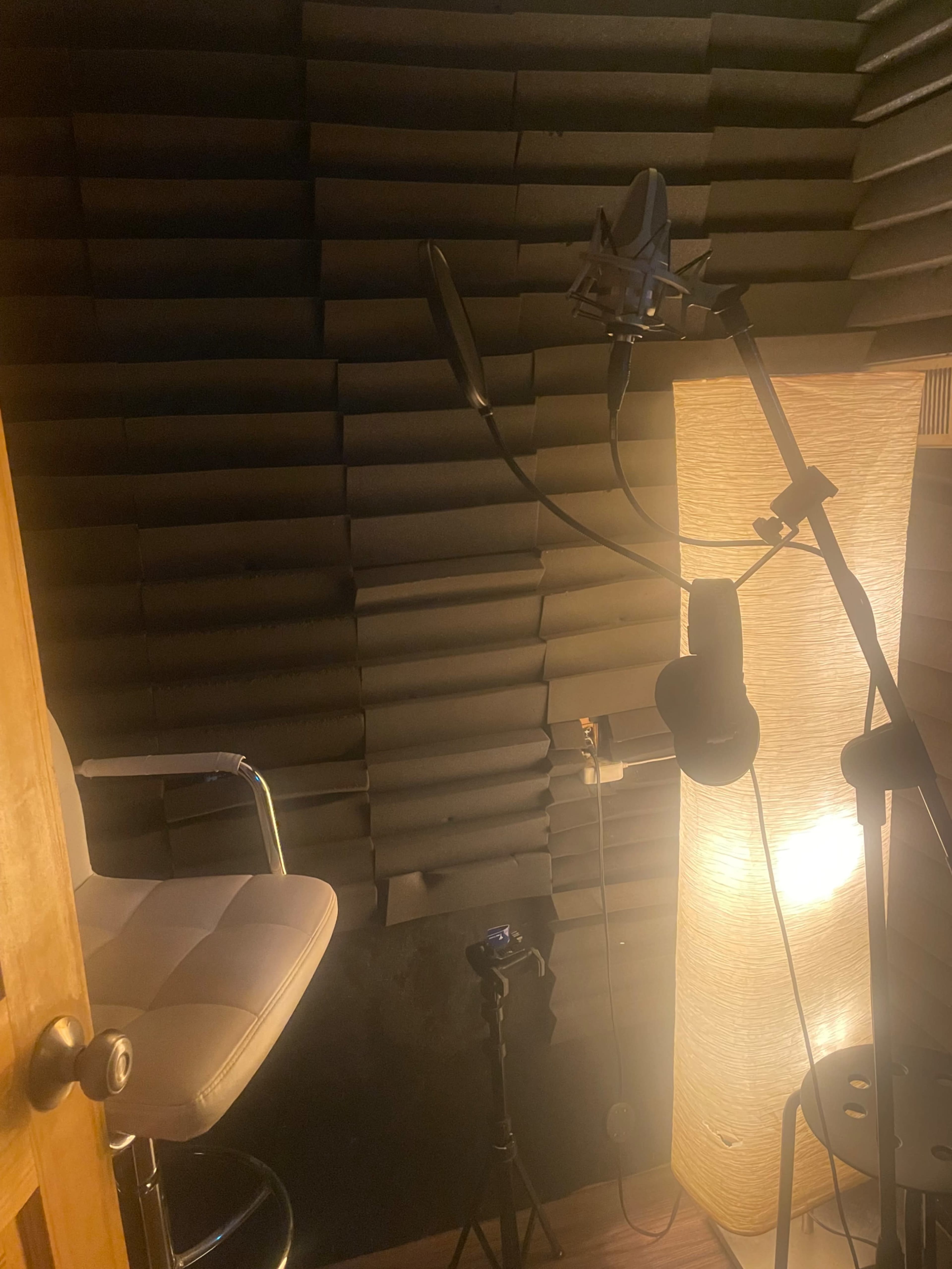The image shows a small recording studio booth with acoustic paneling, a microphone on a stand, a stool, and a lamp providing soft lighting.
