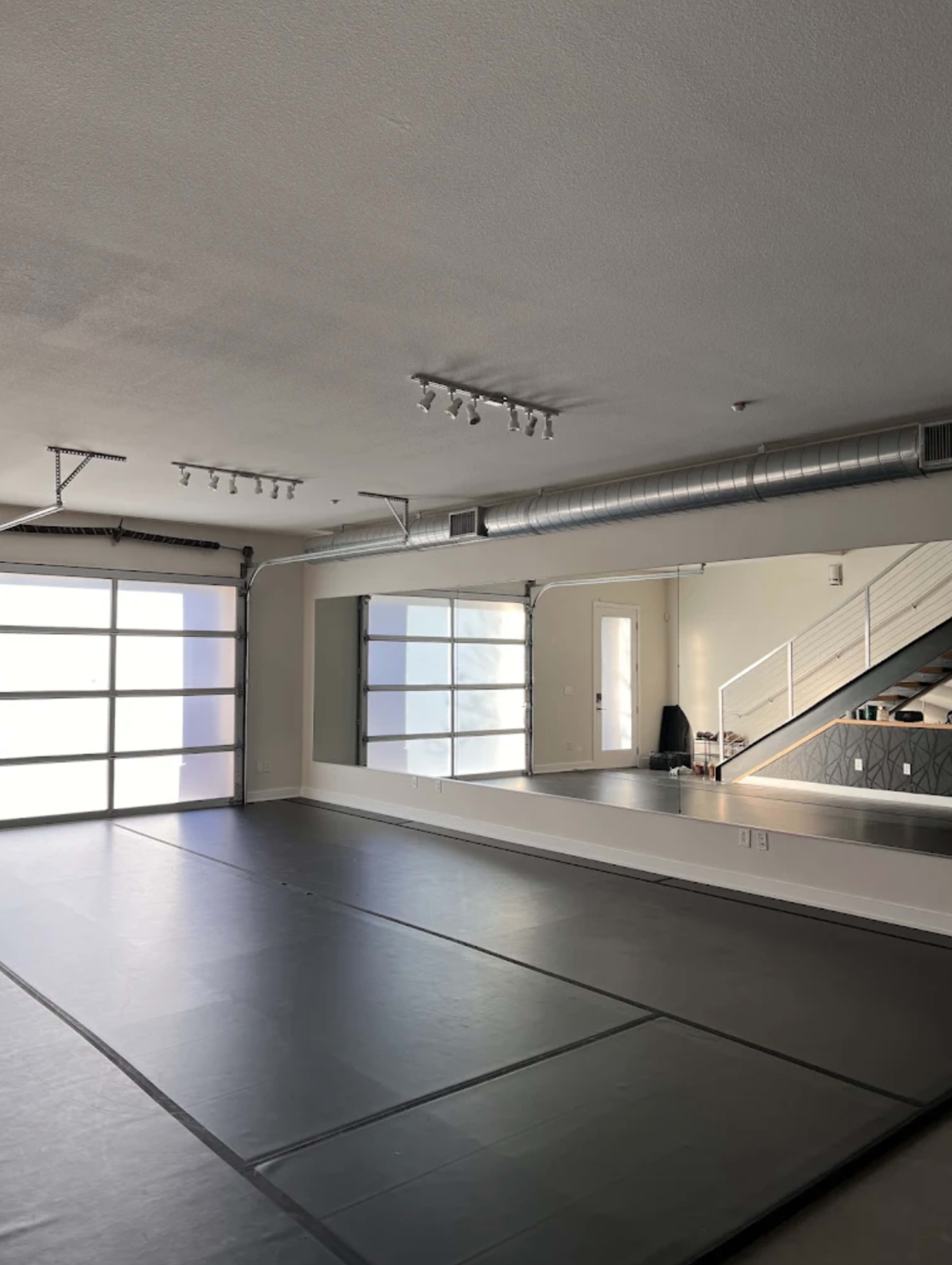 The image shows an empty gym or studio space with a mirrored wall and large glass garage door, illuminated by overhead lighting.