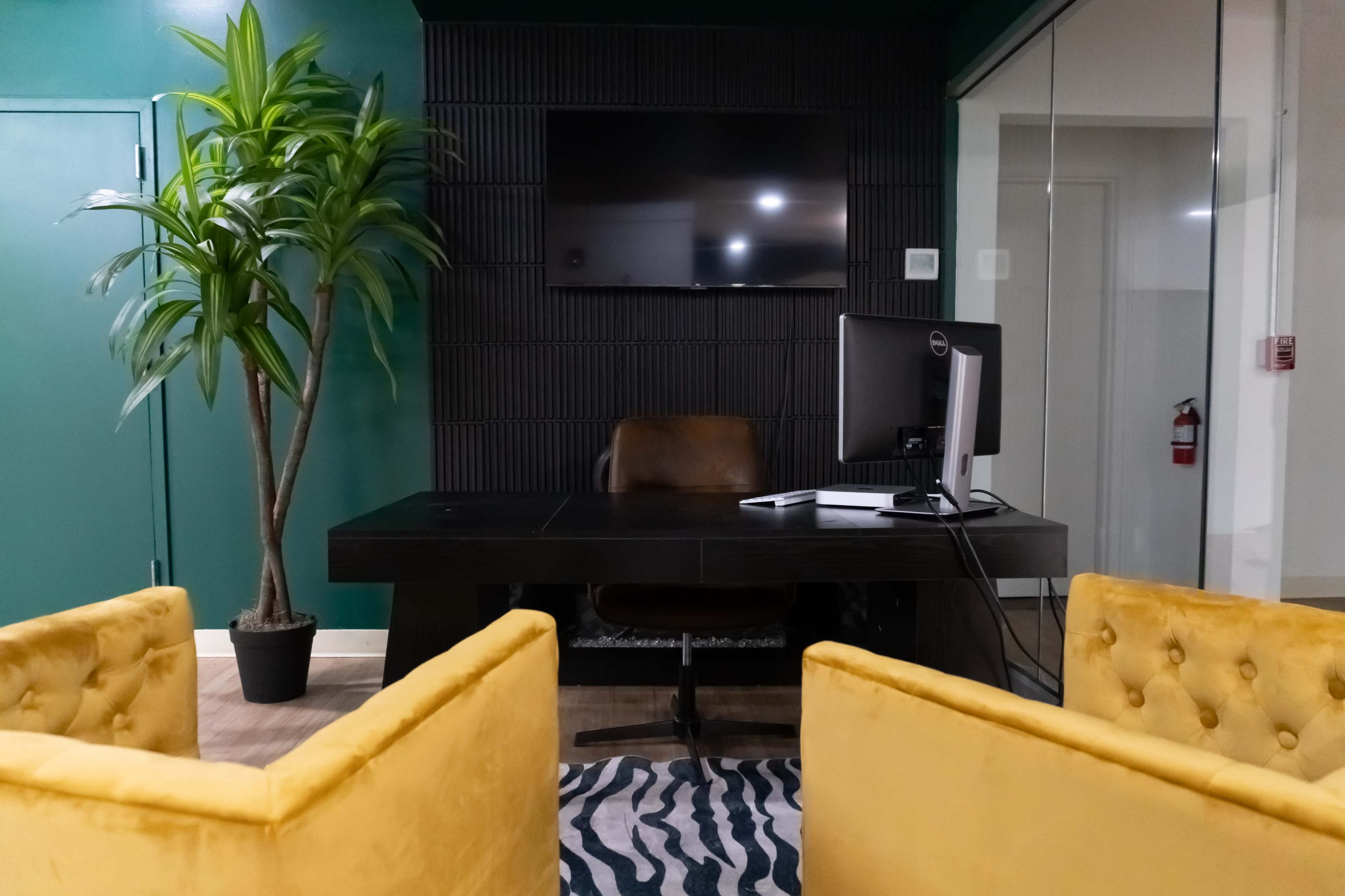 A modern office space features a desk with a computer, a leather chair, two yellow upholstered chairs, a large television mounted on the wall, and a potted plant.