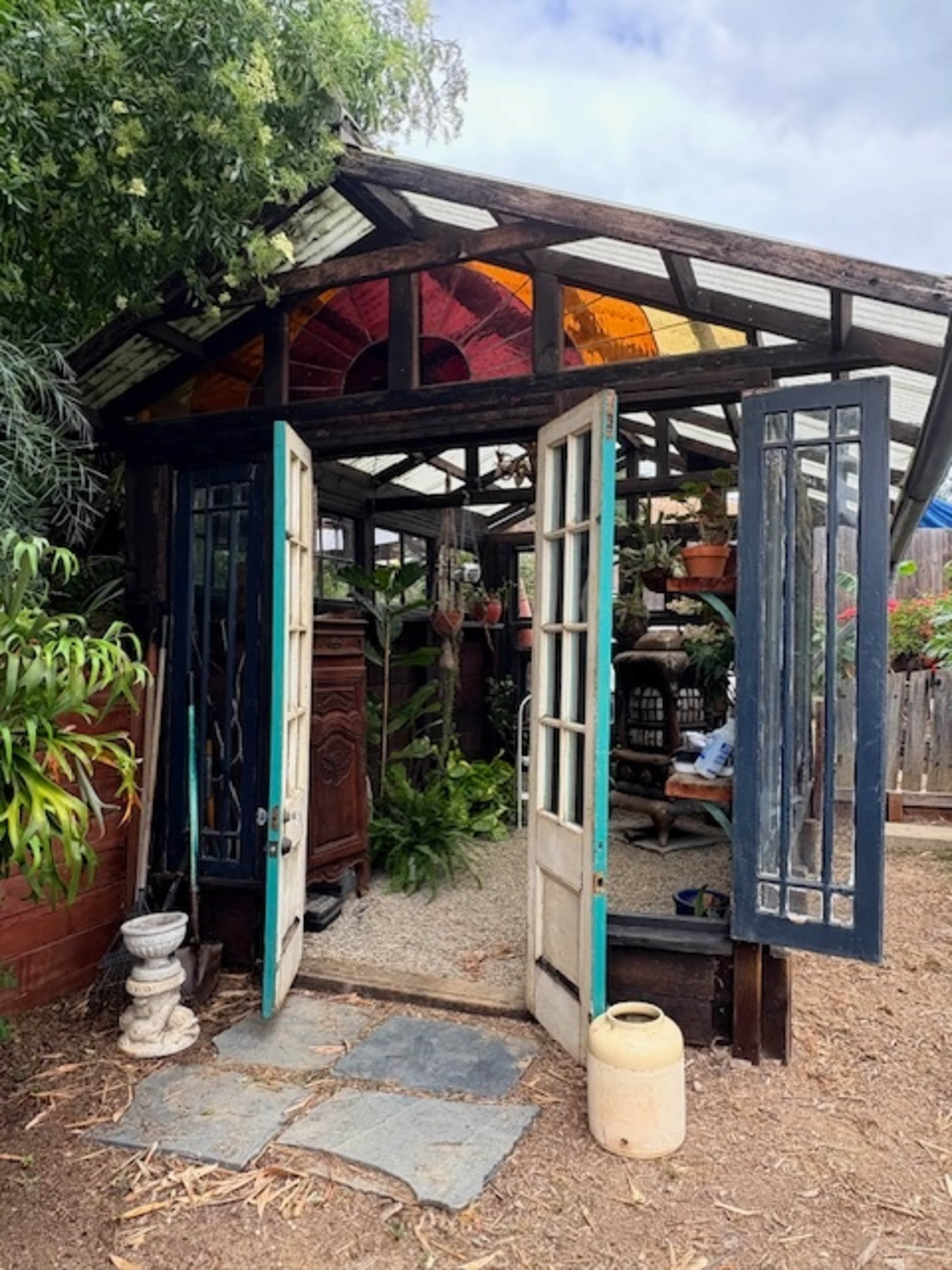 A rustic greenhouse features open double doors with stained glass accents and is surrounded by various plants.