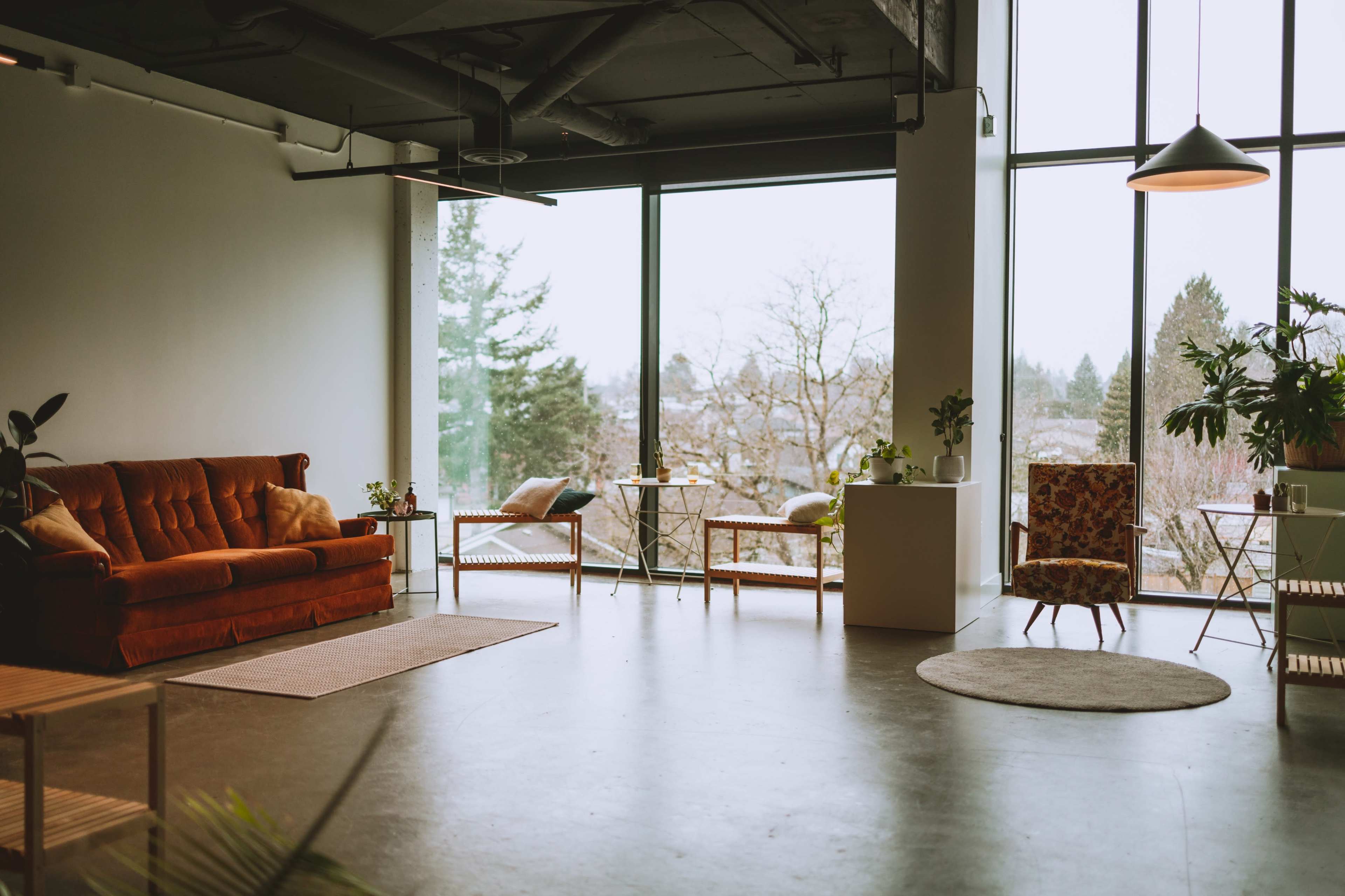 A spacious, modern living area with large windows, a rust-colored sofa, and various potted plants.