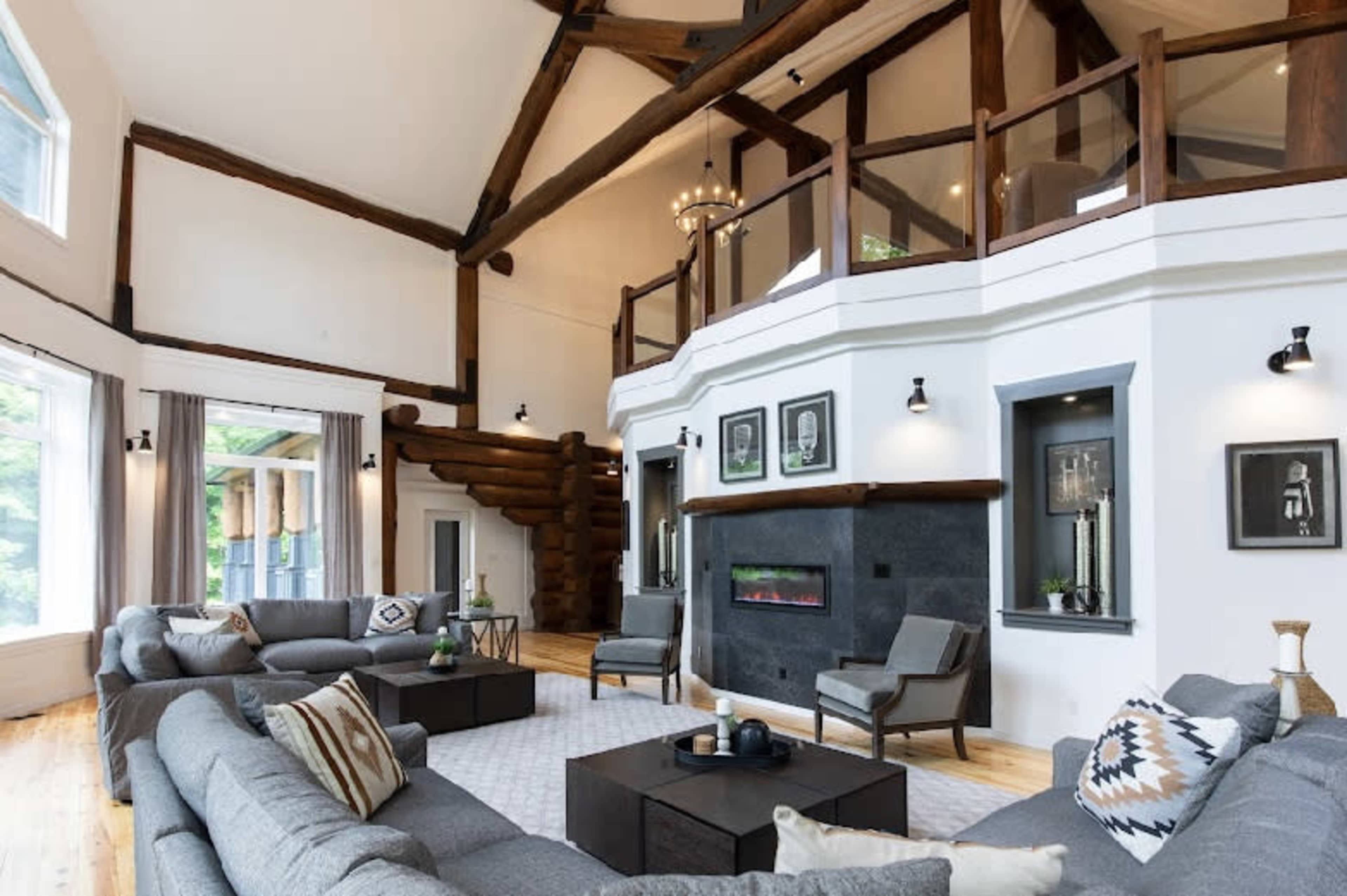 A spacious living room features high ceilings with exposed wooden beams, large windows, and modern furniture arranged around a central coffee table.