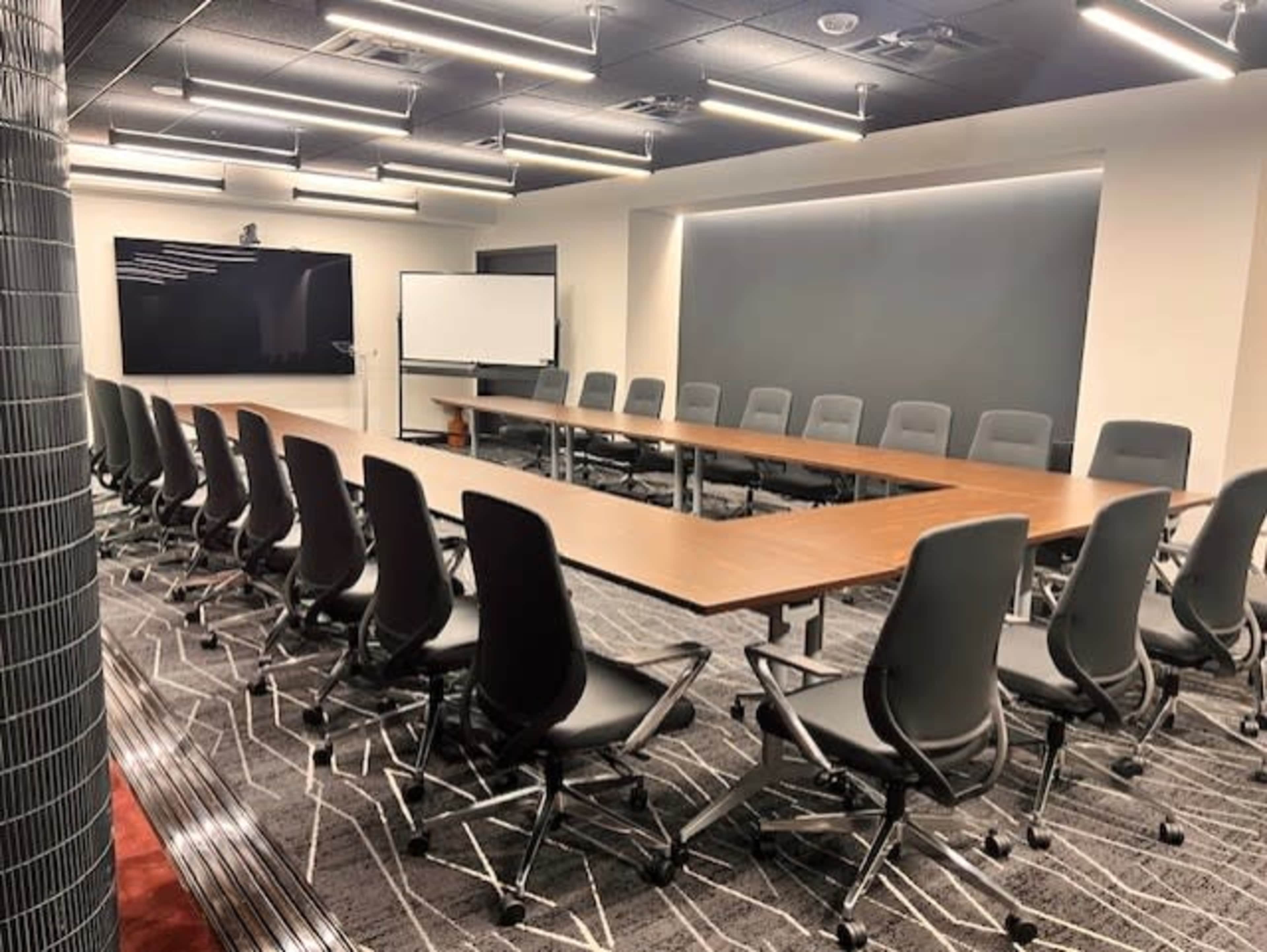 The image shows a modern conference room with a long, U-shaped table surrounded by ergonomic chairs, equipped with multiple screens for presentations.
