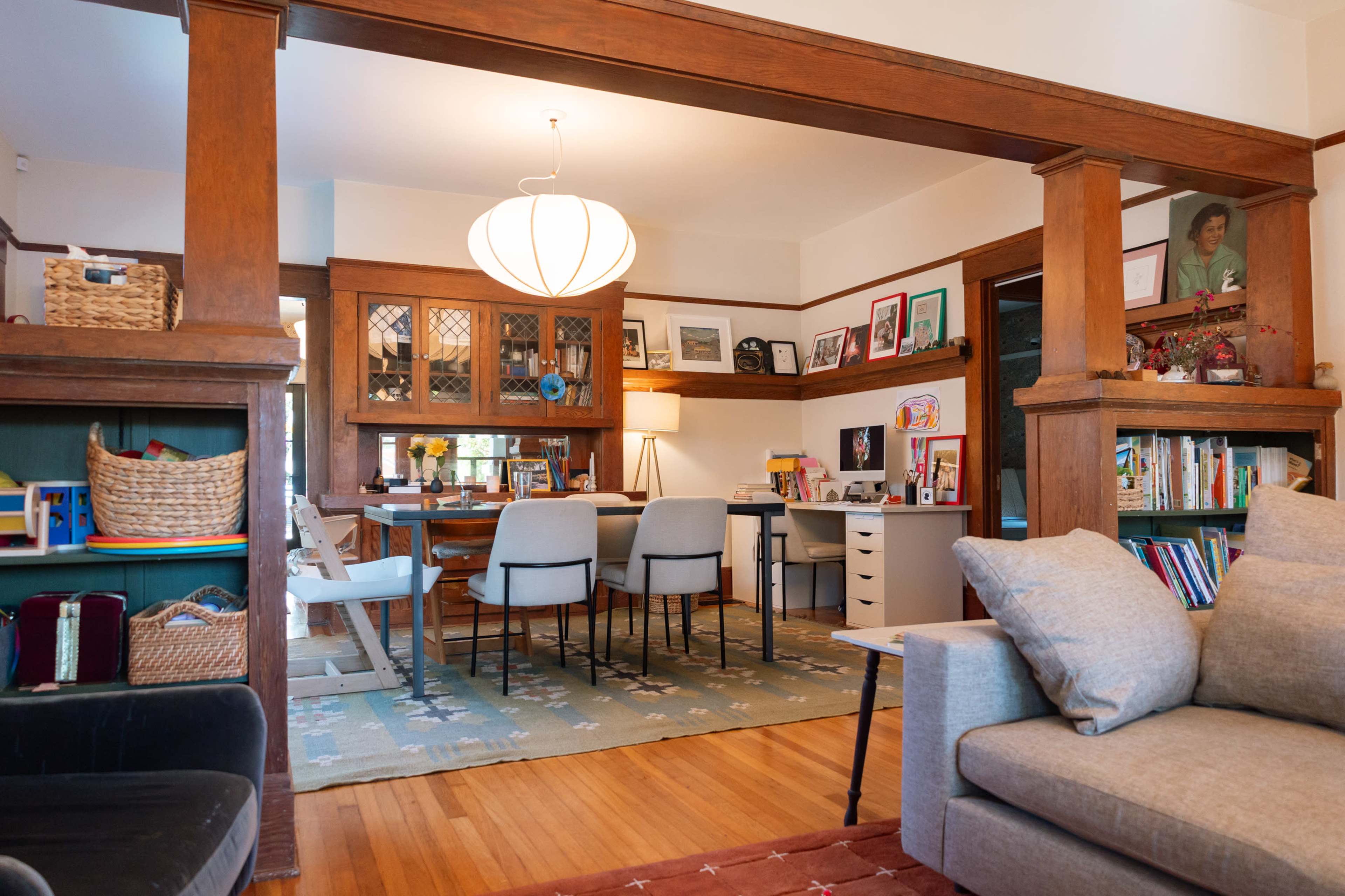 The image shows a cozy living room with a view into a well-lit home office area featuring a modern desk, bookshelves, and decorative artwork.