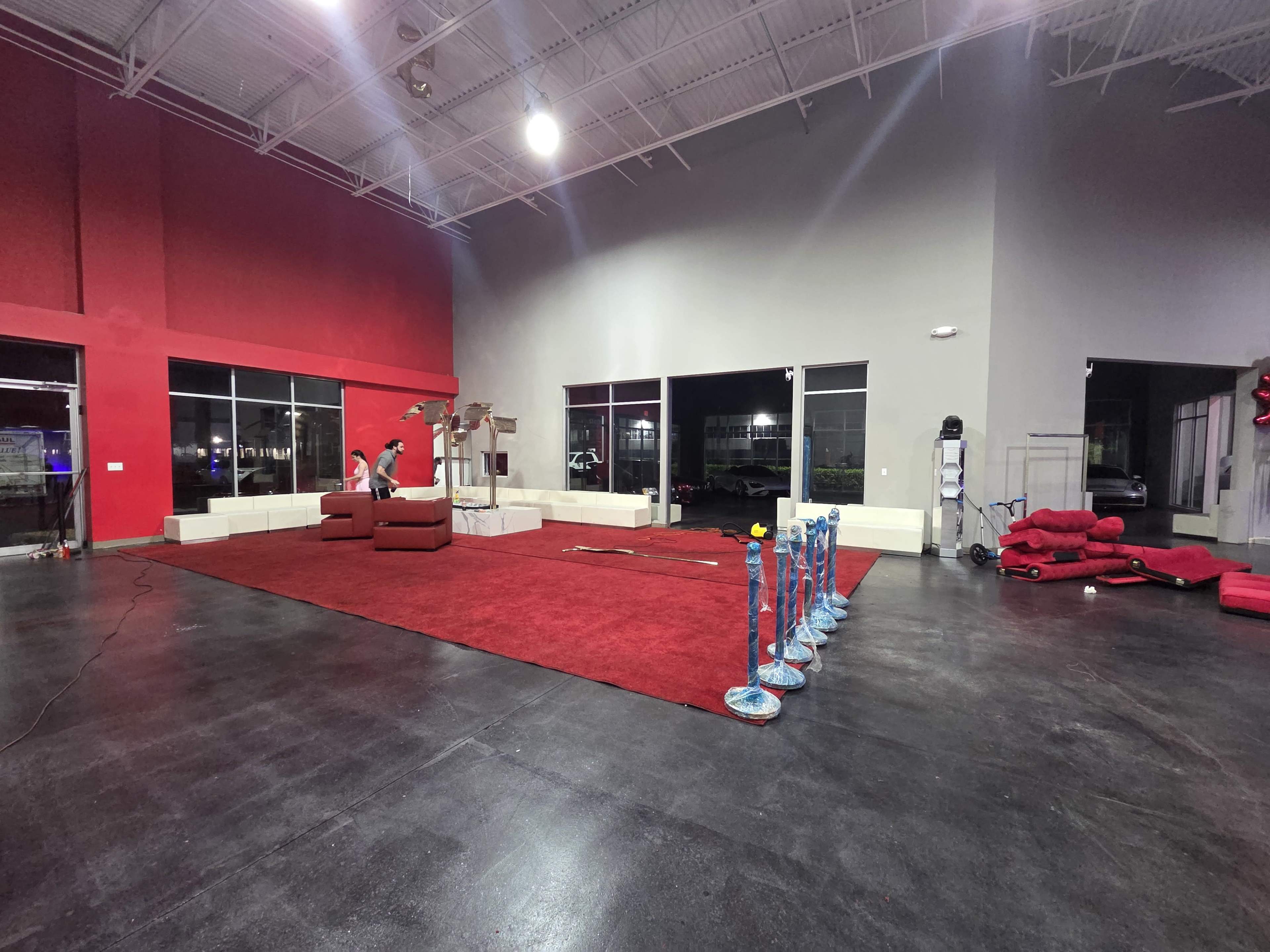 The image shows an empty commercial space with a large red carpet, a few red couches, and blue stanchions lined up in front of the walls, while a person is seen working in the background.