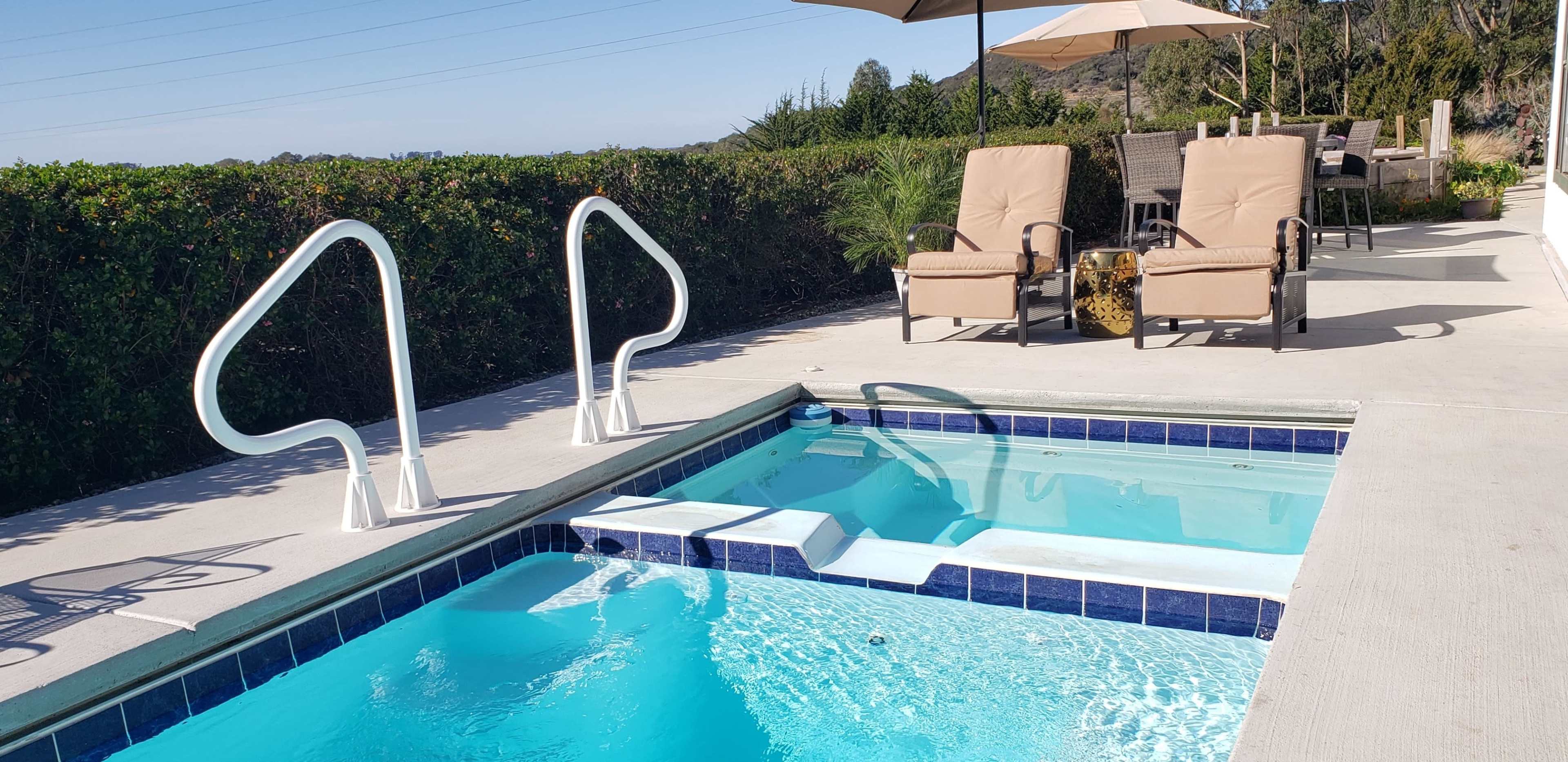 A swimming pool with blue water, a set of steps, and two lounge chairs on a patio beside it.