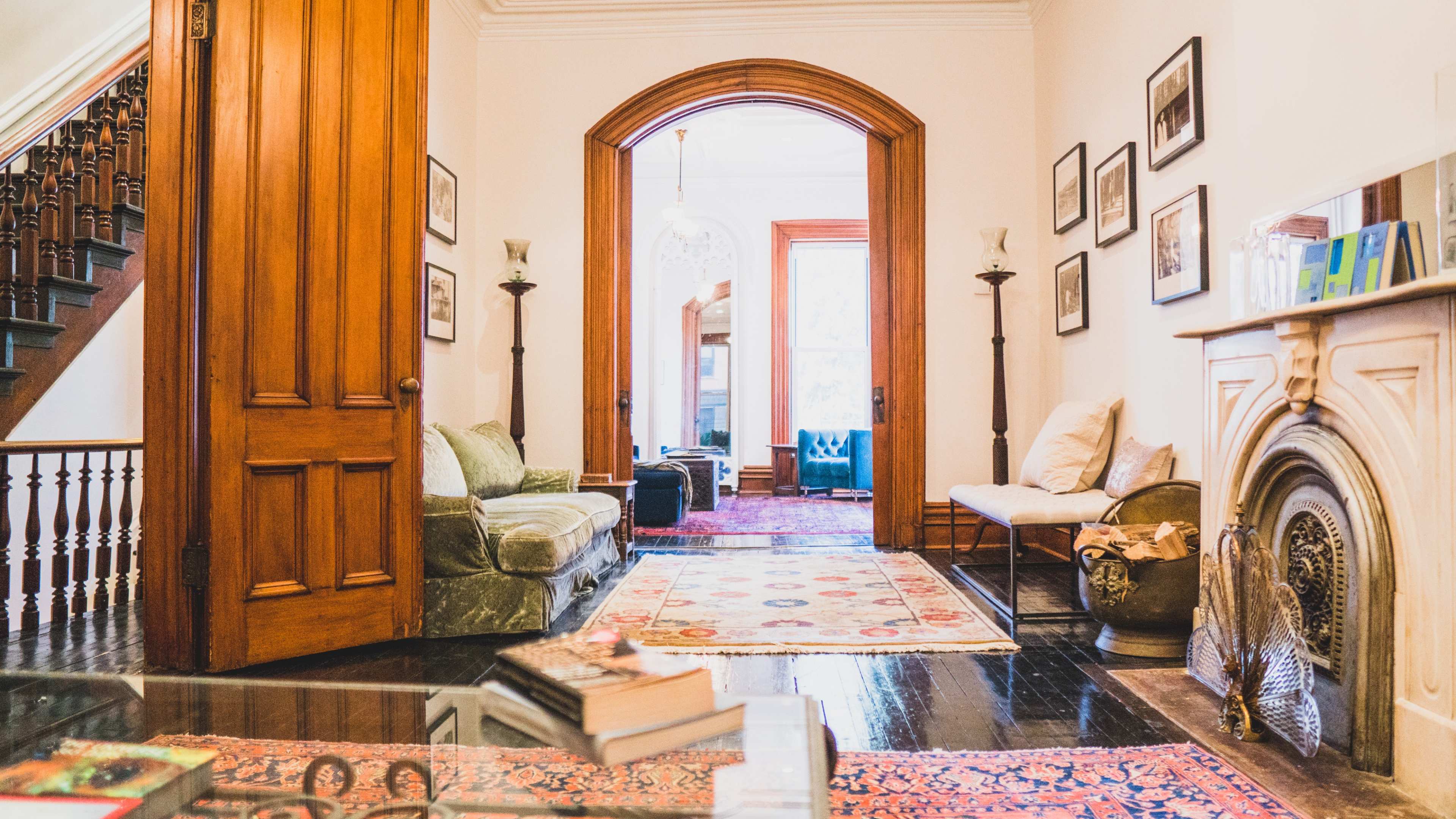 The image shows a warm interior of a home with wooden doors, a staircase, a plush sofa, and an ornate fireplace, all adorned with framed photographs and a patterned rug.