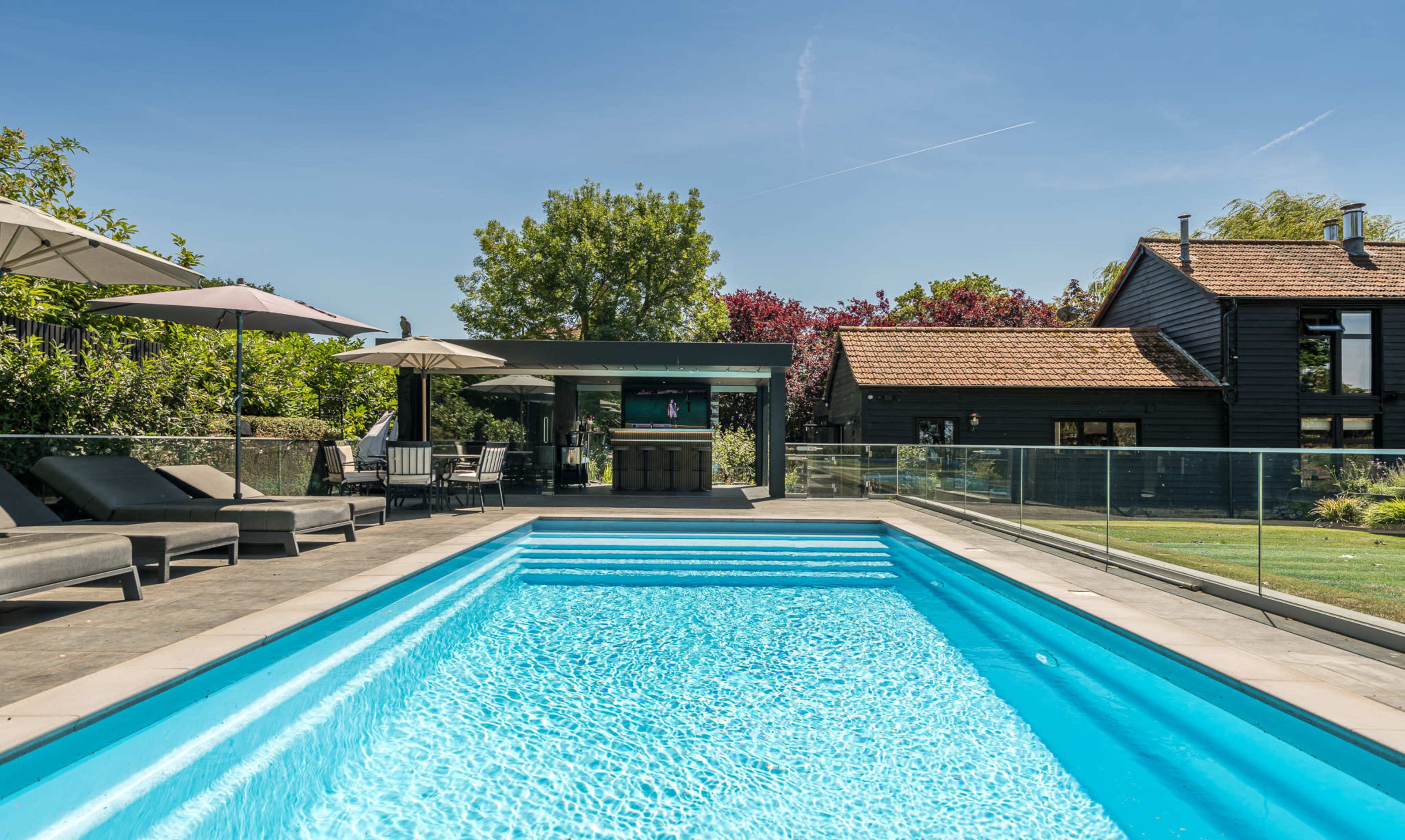 The image shows a modern swimming pool surrounded by lounge chairs and umbrellas, with greenery and a house in the background.