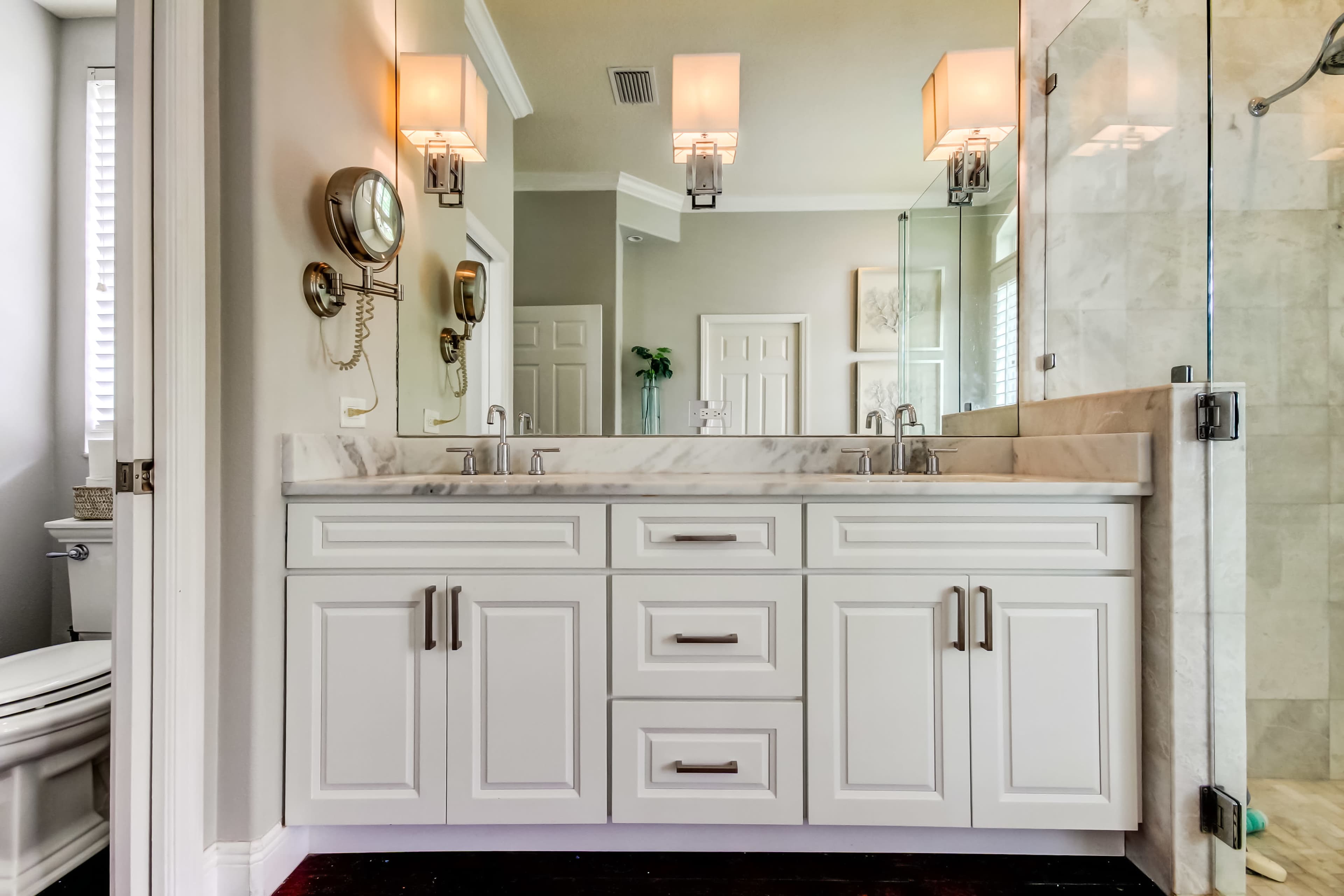 A modern bathroom features a double sink vanity with a marble countertop, two wall-mounted light fixtures, and a glass shower enclosure.