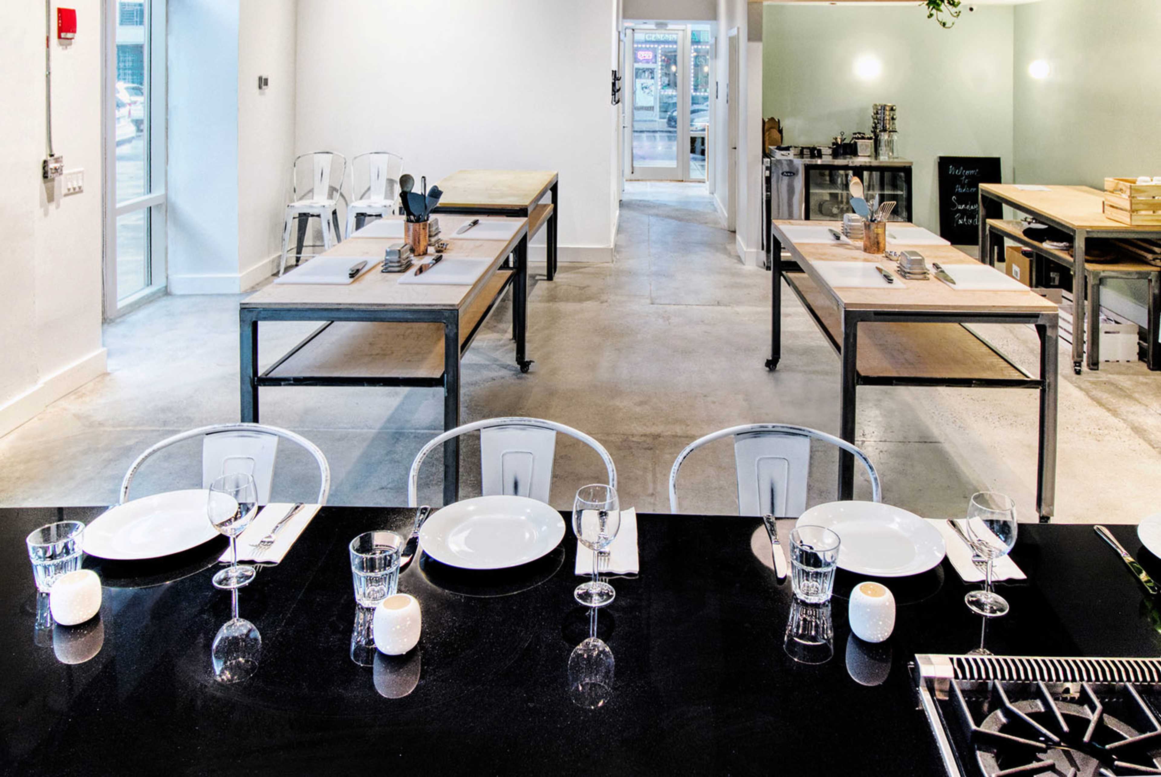 A modern kitchen space with several dining tables, neatly set with plates and utensils, and a view of a bright hallway in the background.