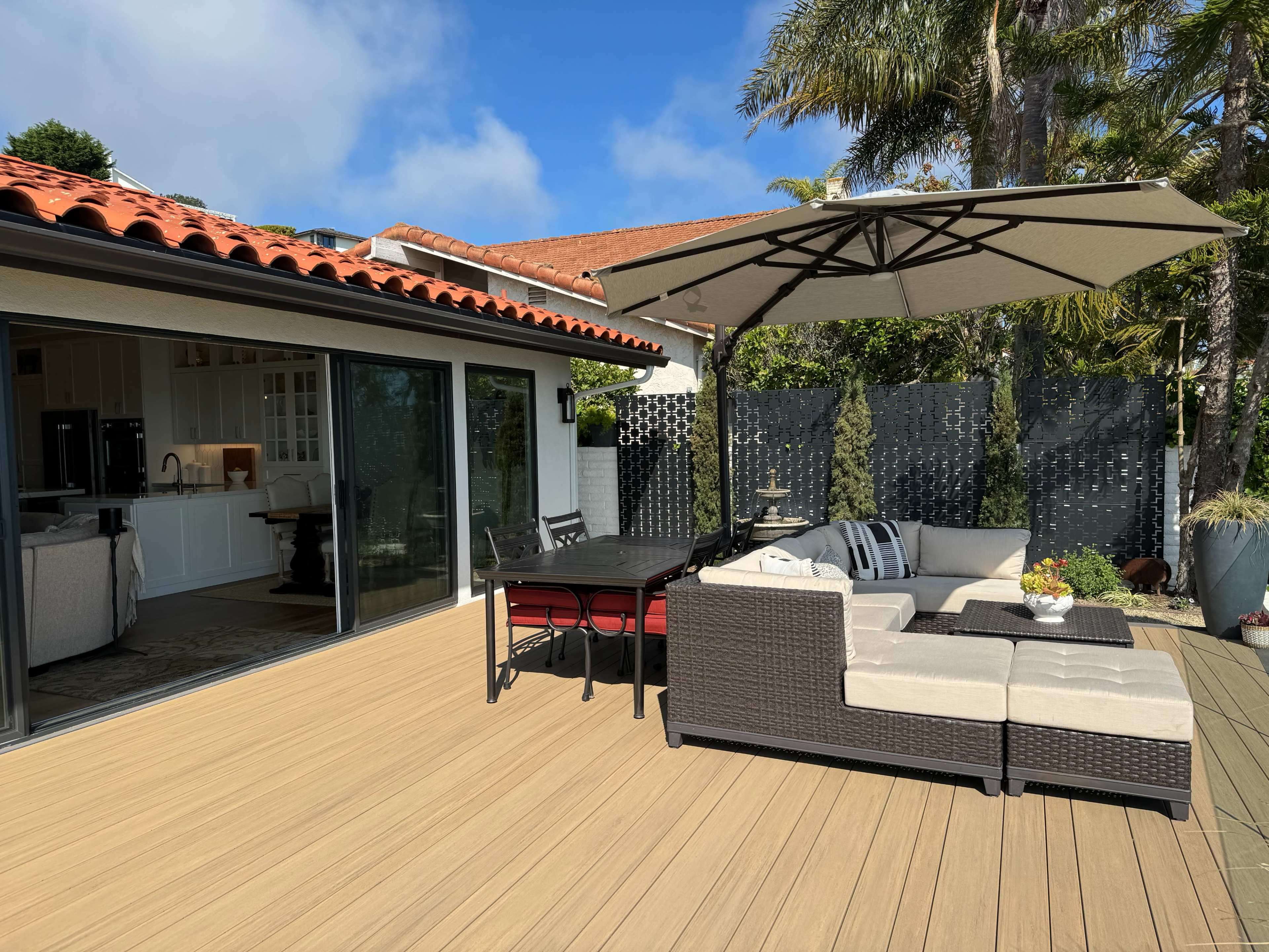 The image shows a patio area with a seating arrangement and an umbrella beside a house with a kitchen visible through large glass doors.