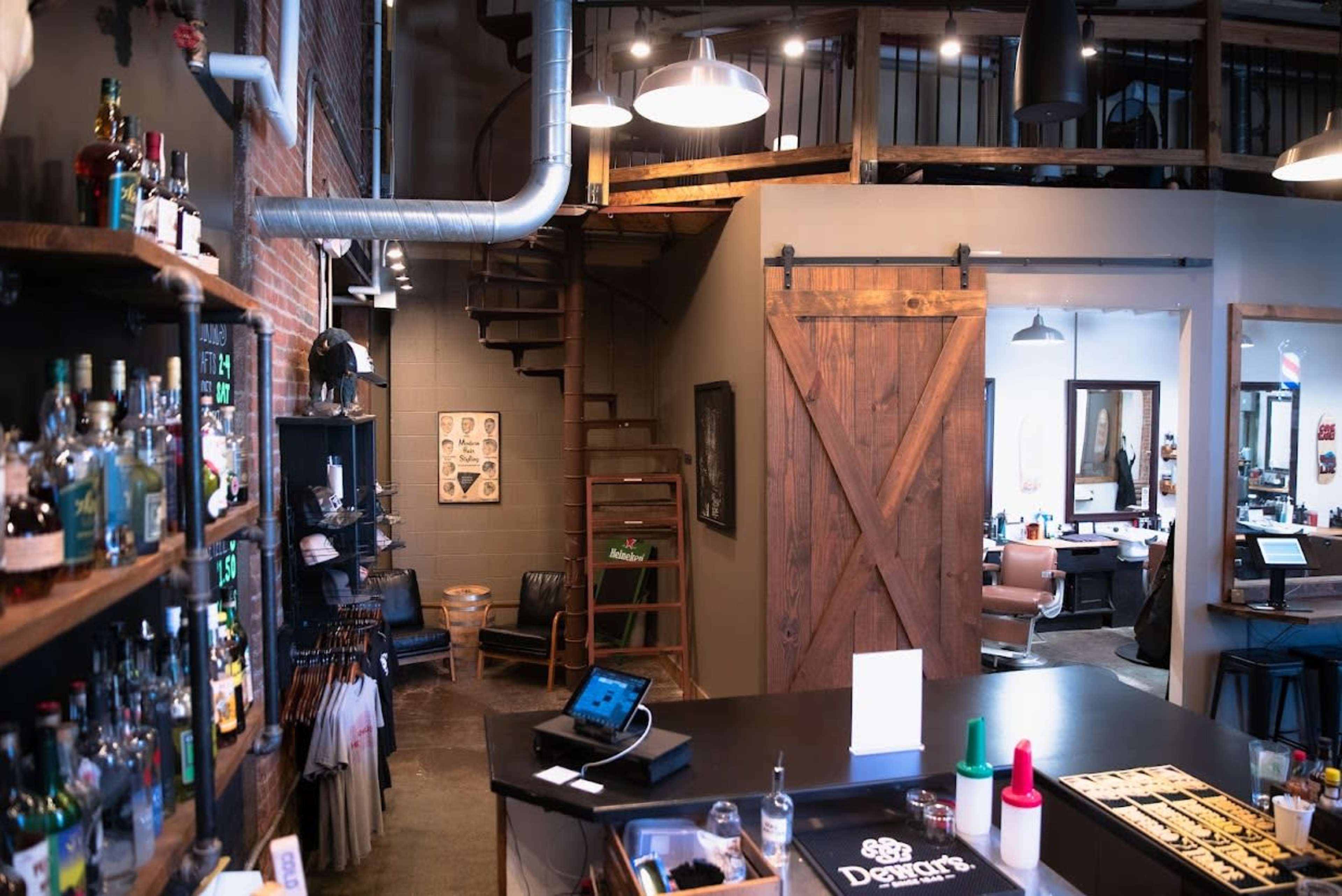 The image shows a barbershop interior featuring a wooden counter, shelves with various bottles, and a visible spiral staircase leading to an upper level.