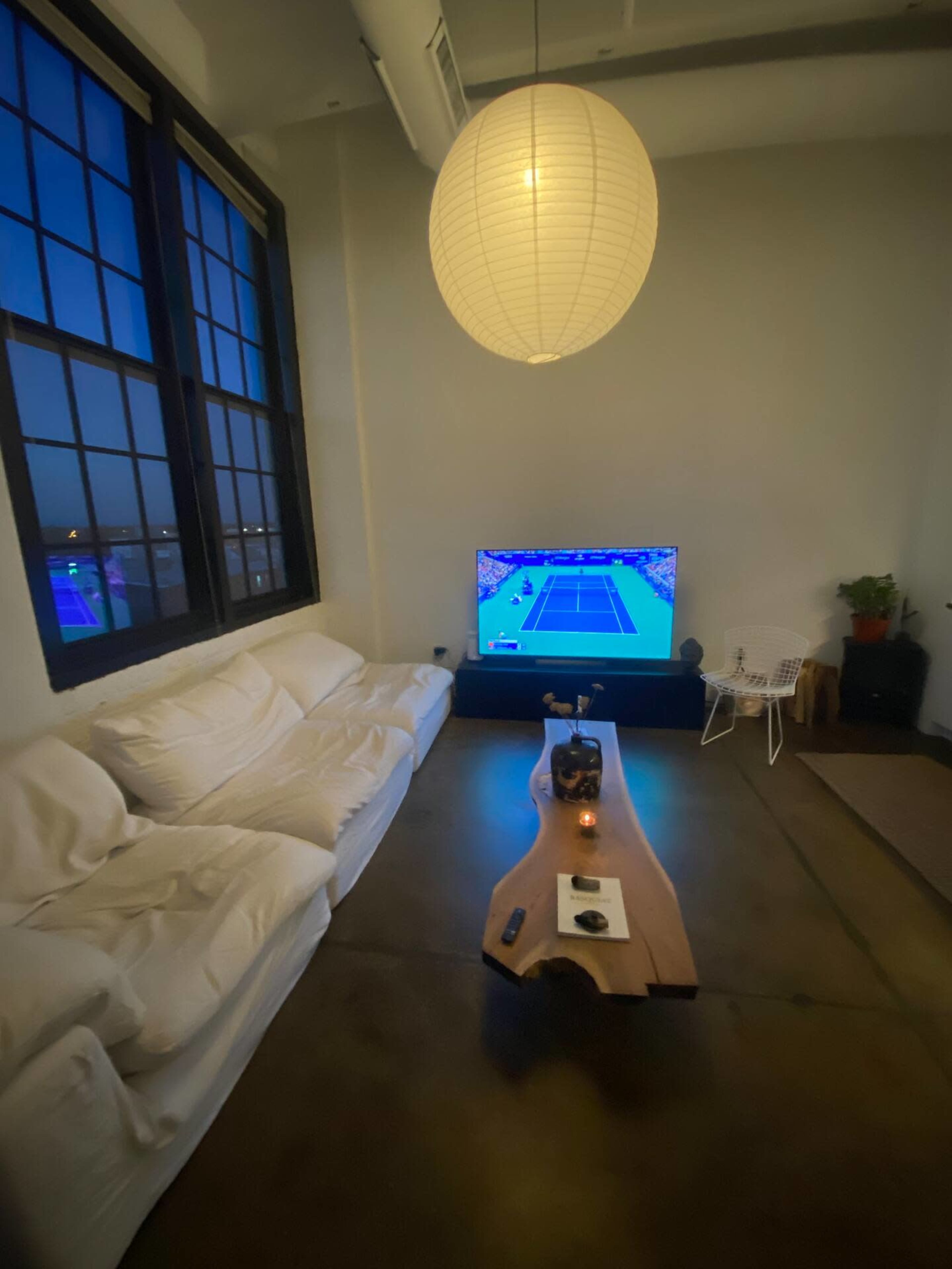 A cozy living room features a large white sofa, a wooden coffee table with decorative items, a television displaying a tennis match, and a large window letting in evening light.