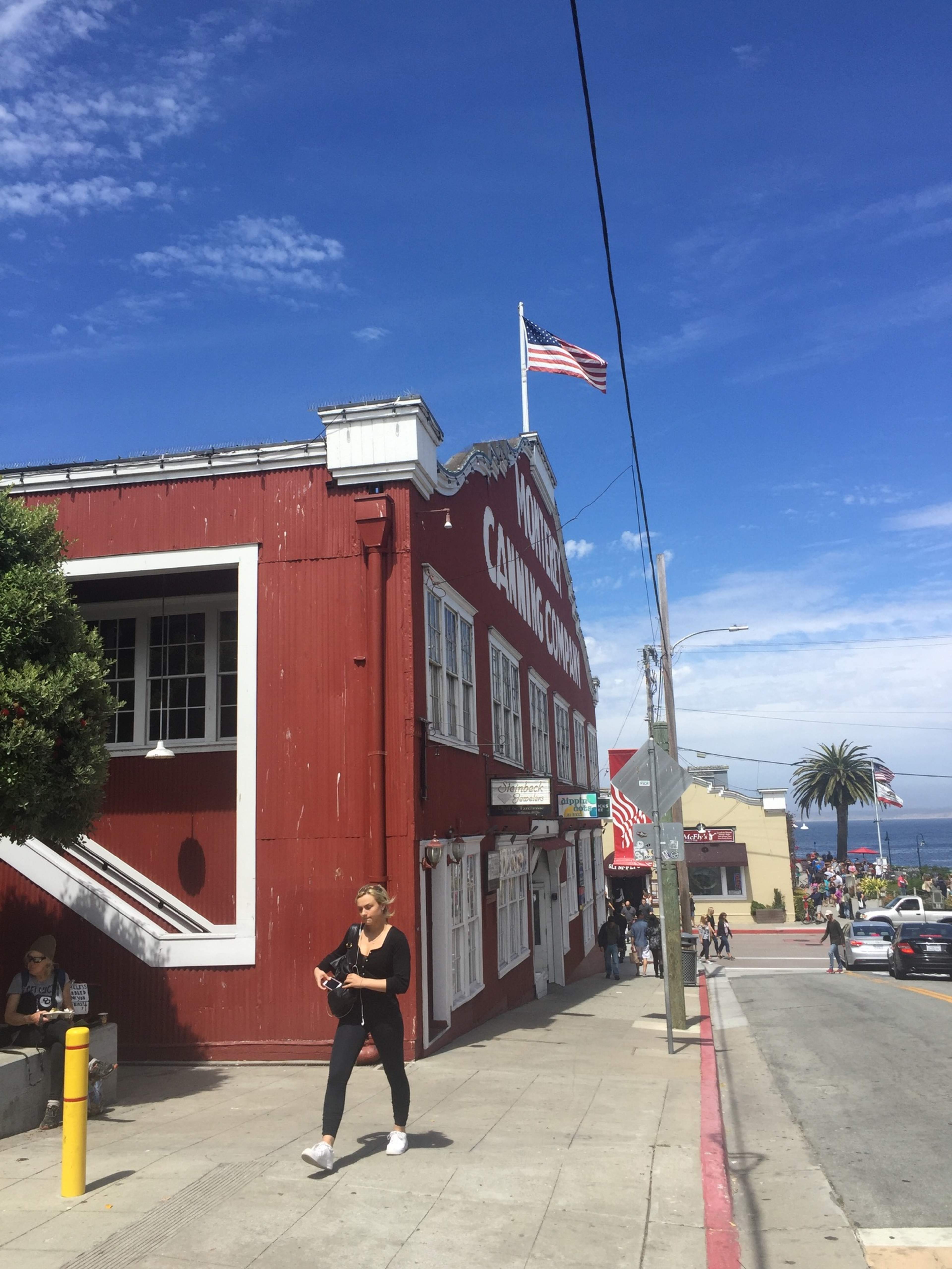 A woman walks past a red building marked "Monterey Canning Company" with an American flag flying above, while the ocean is visible in the background.