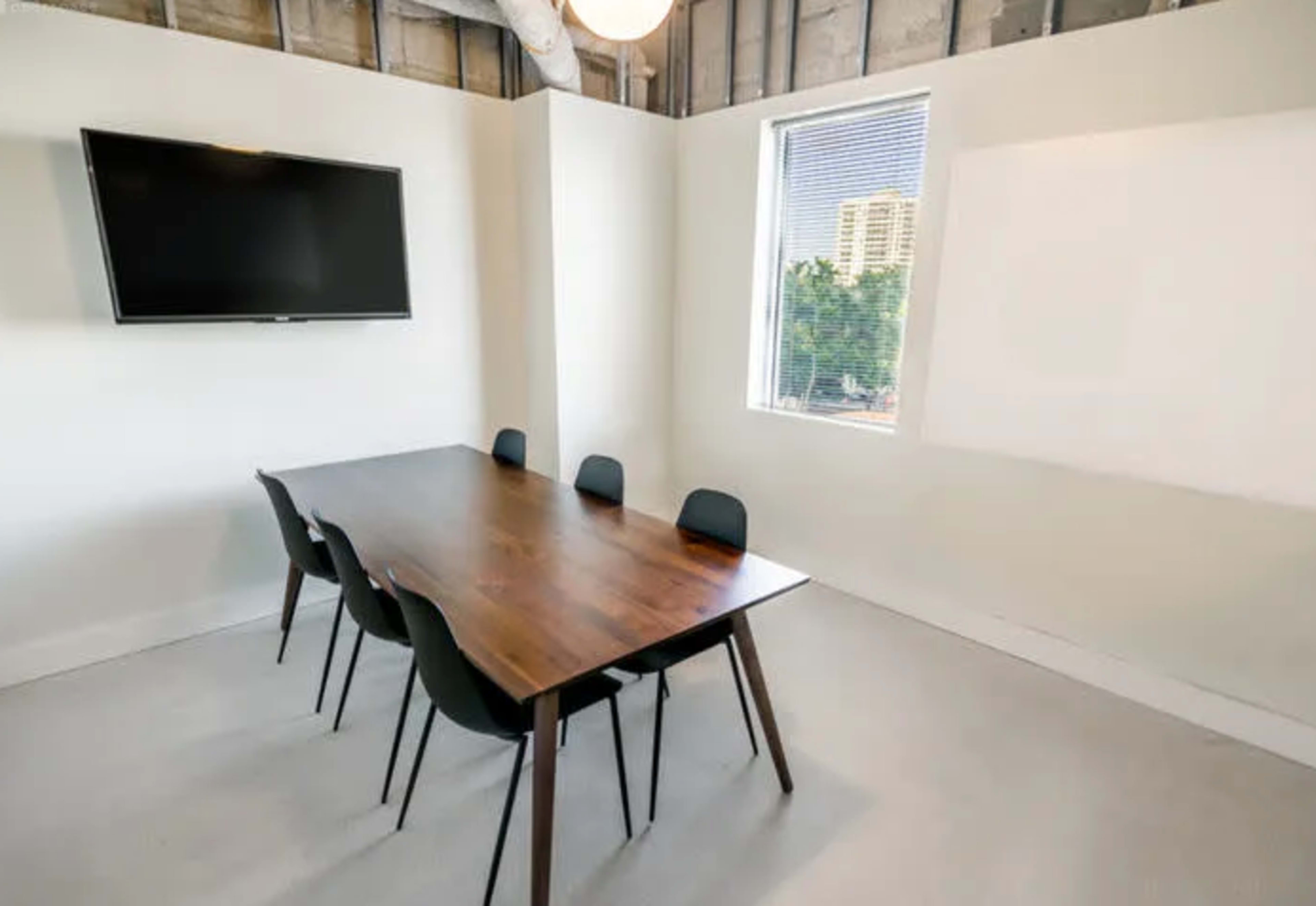A conference room features a wooden table surrounded by six black chairs, with a wall-mounted television and a window displaying city views.