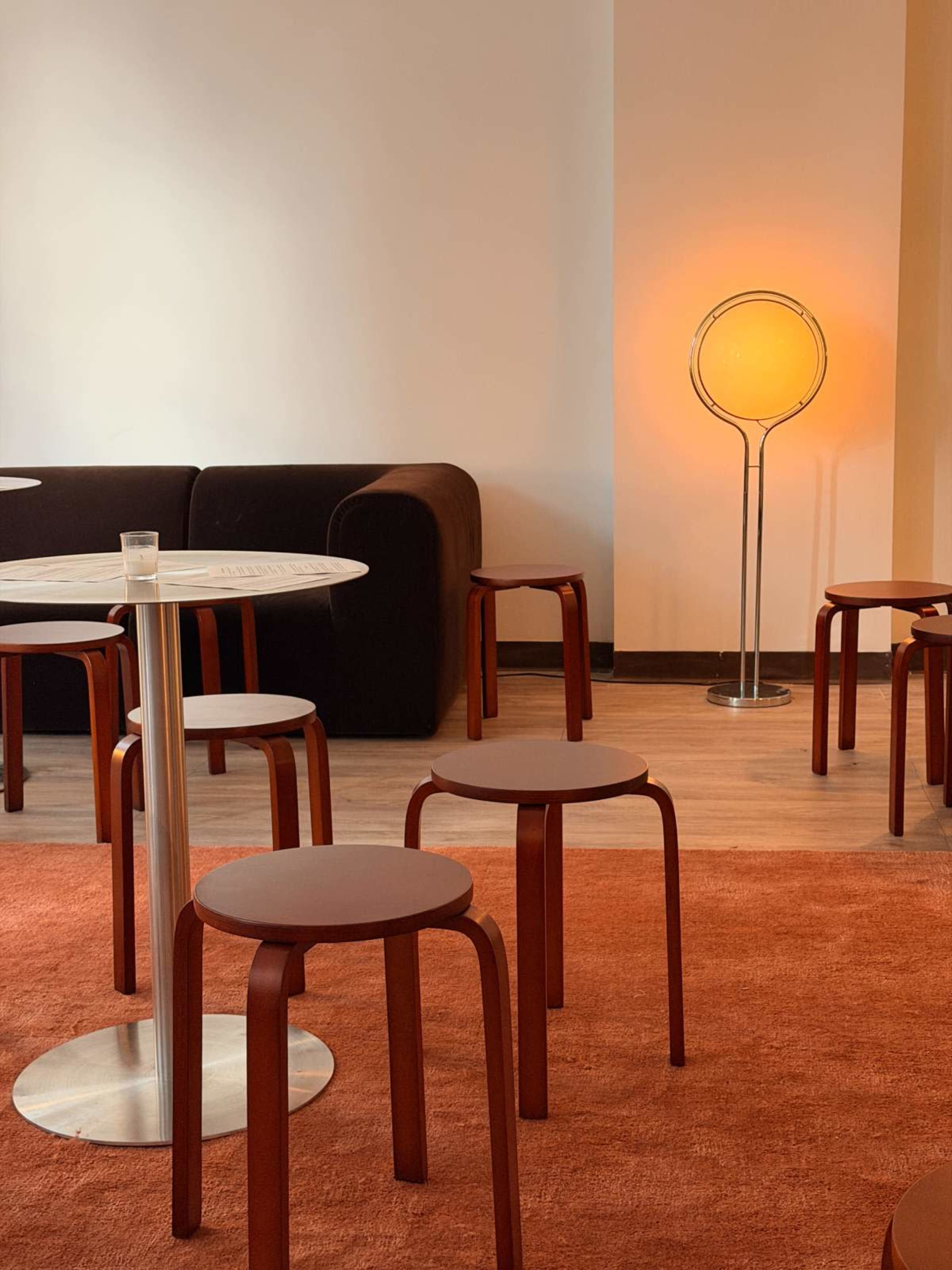 A minimalist interior features a round table with a glass on top, surrounded by red stools and a floor lamp emitting a warm glow.