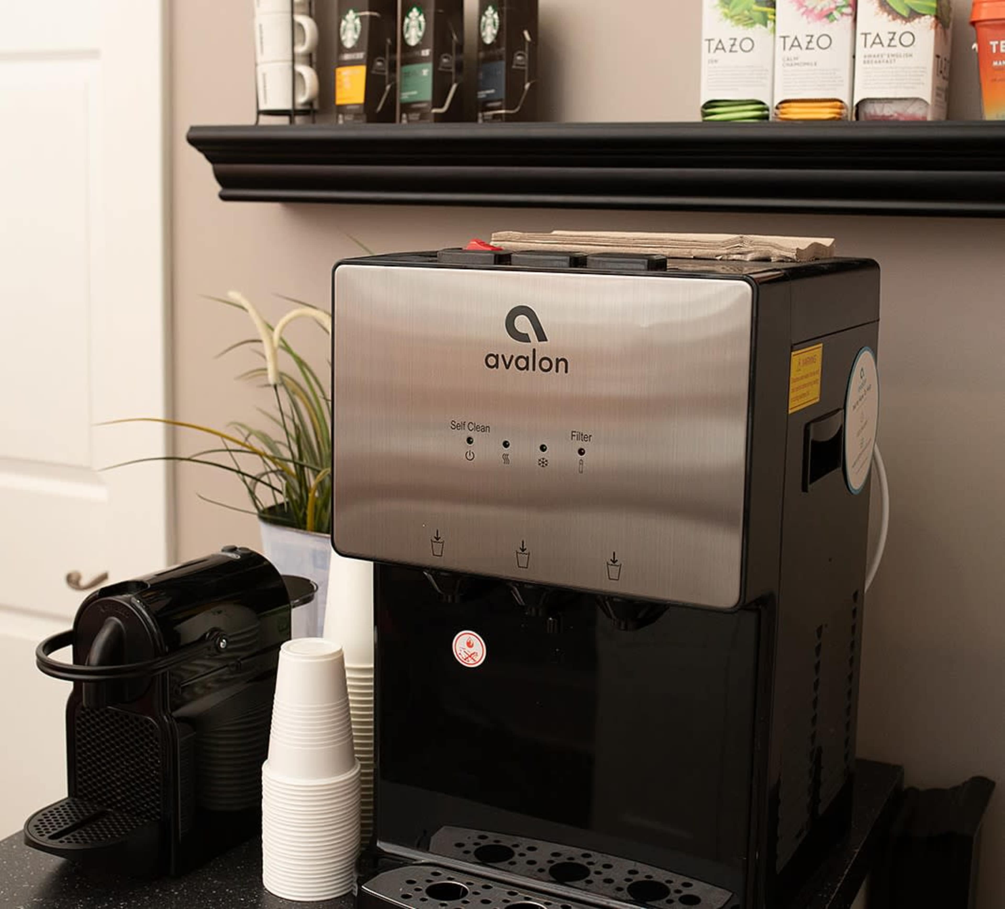 A stainless steel water dispenser with a coffee machine and disposable cups on a countertop.