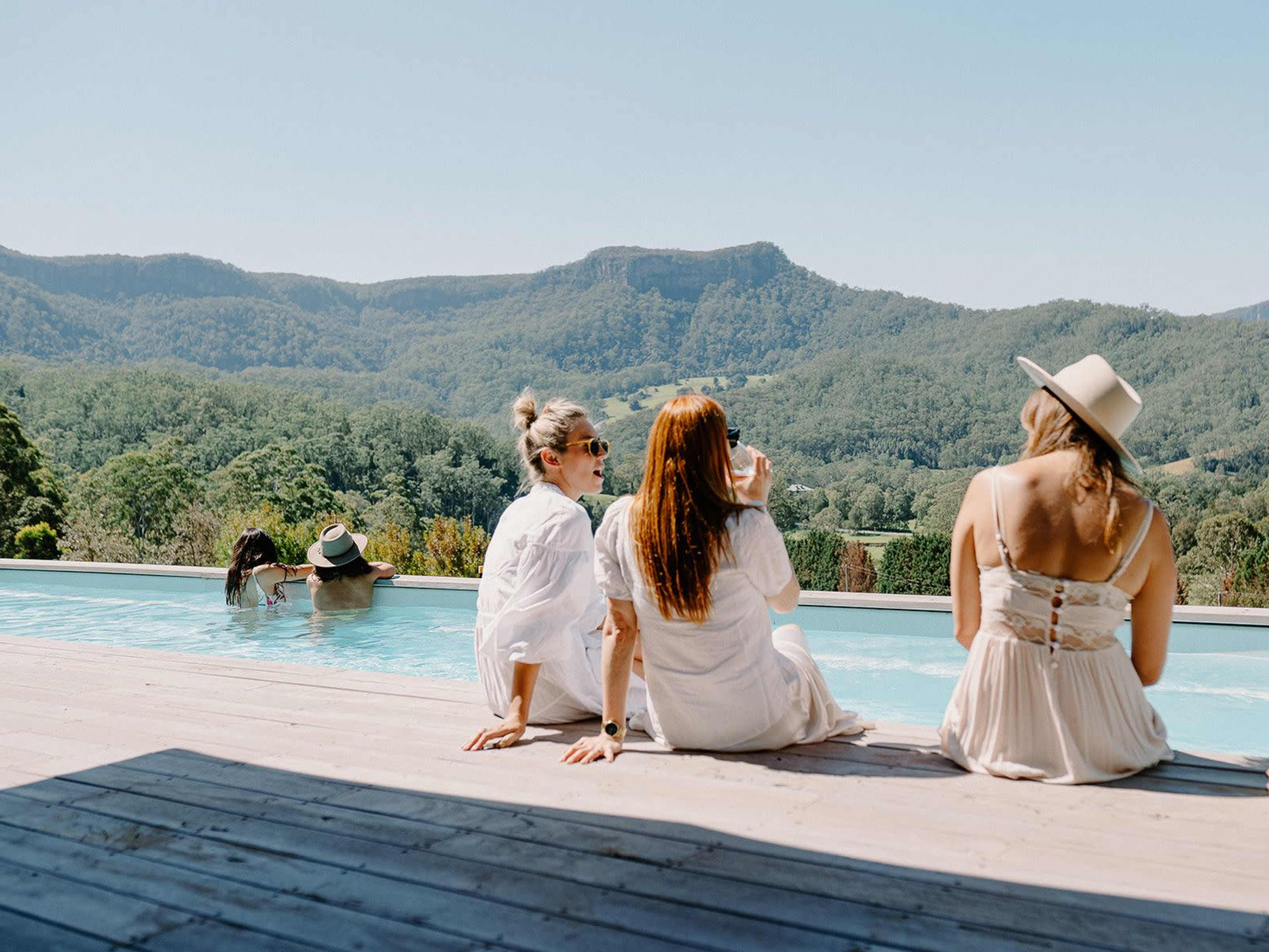 Four women relax by a swimming pool with a scenic mountain backdrop.
