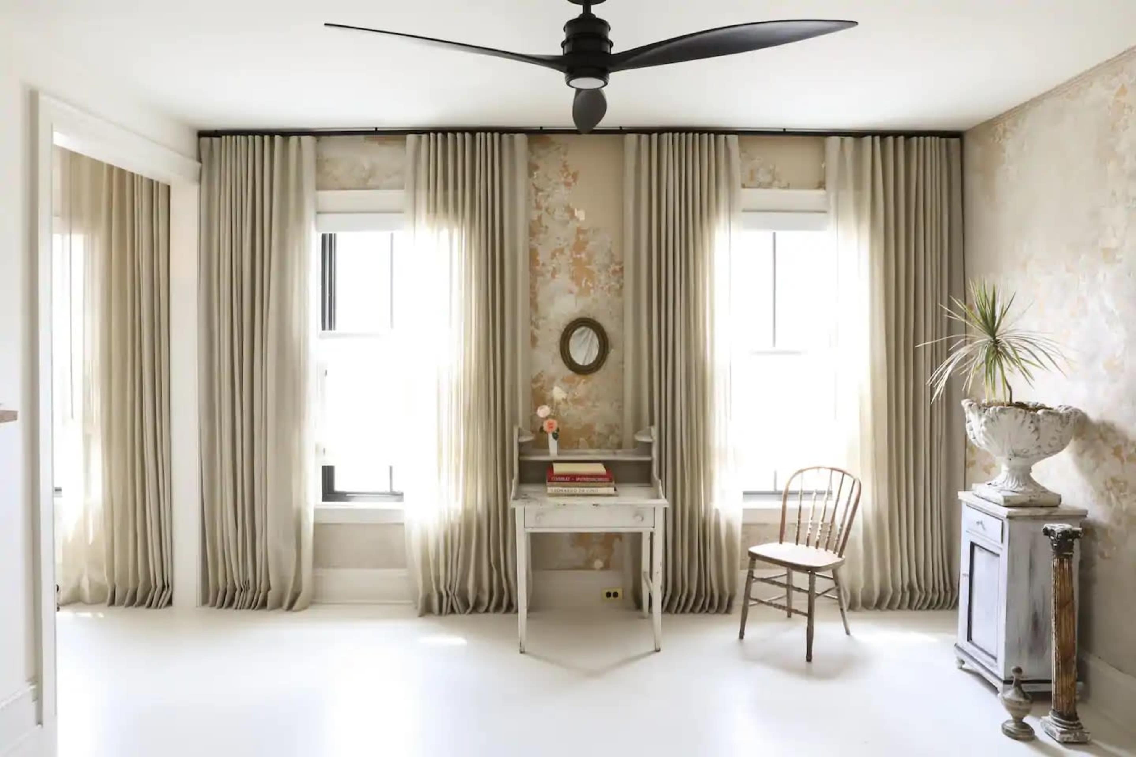 A room features two windows with long curtains, a small desk with books, a wooden chair, and a decorative plant beside an ornate vase.