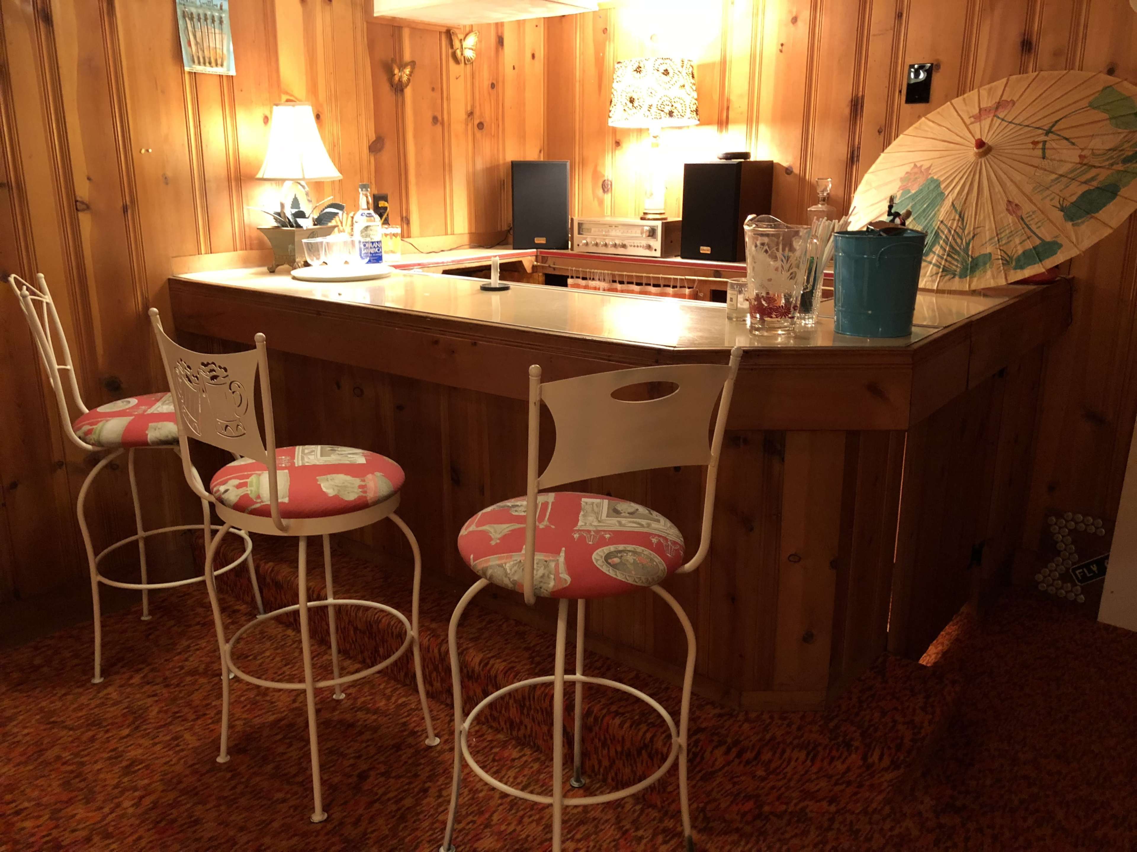 A wooden bar with three metal stools is set against a paneled wall, illuminated by a lamp and decorated with a colorful umbrella.