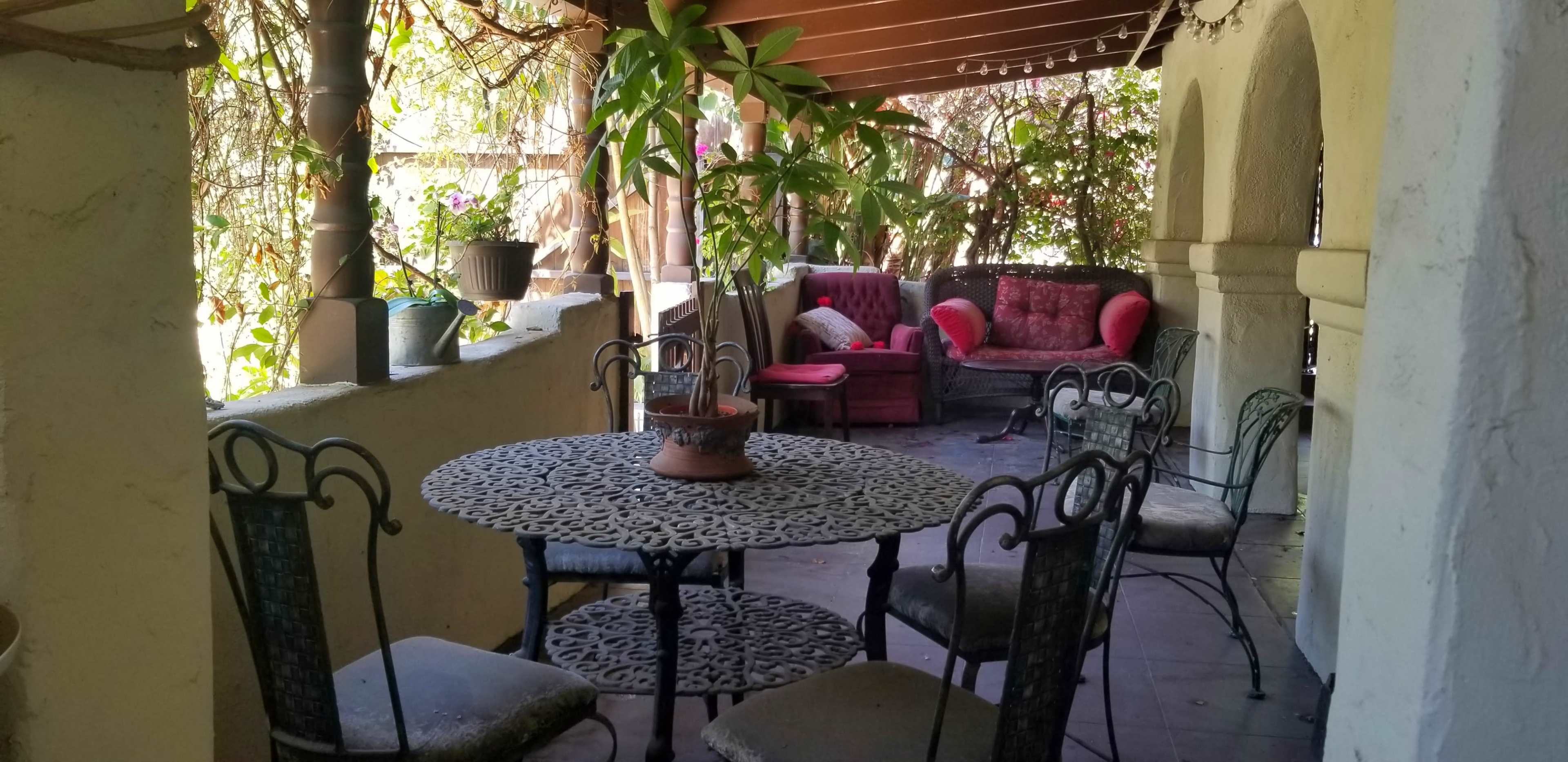 A covered outdoor seating area features a wrought-iron table surrounded by six chairs, with plants and comfortable couches in the background.