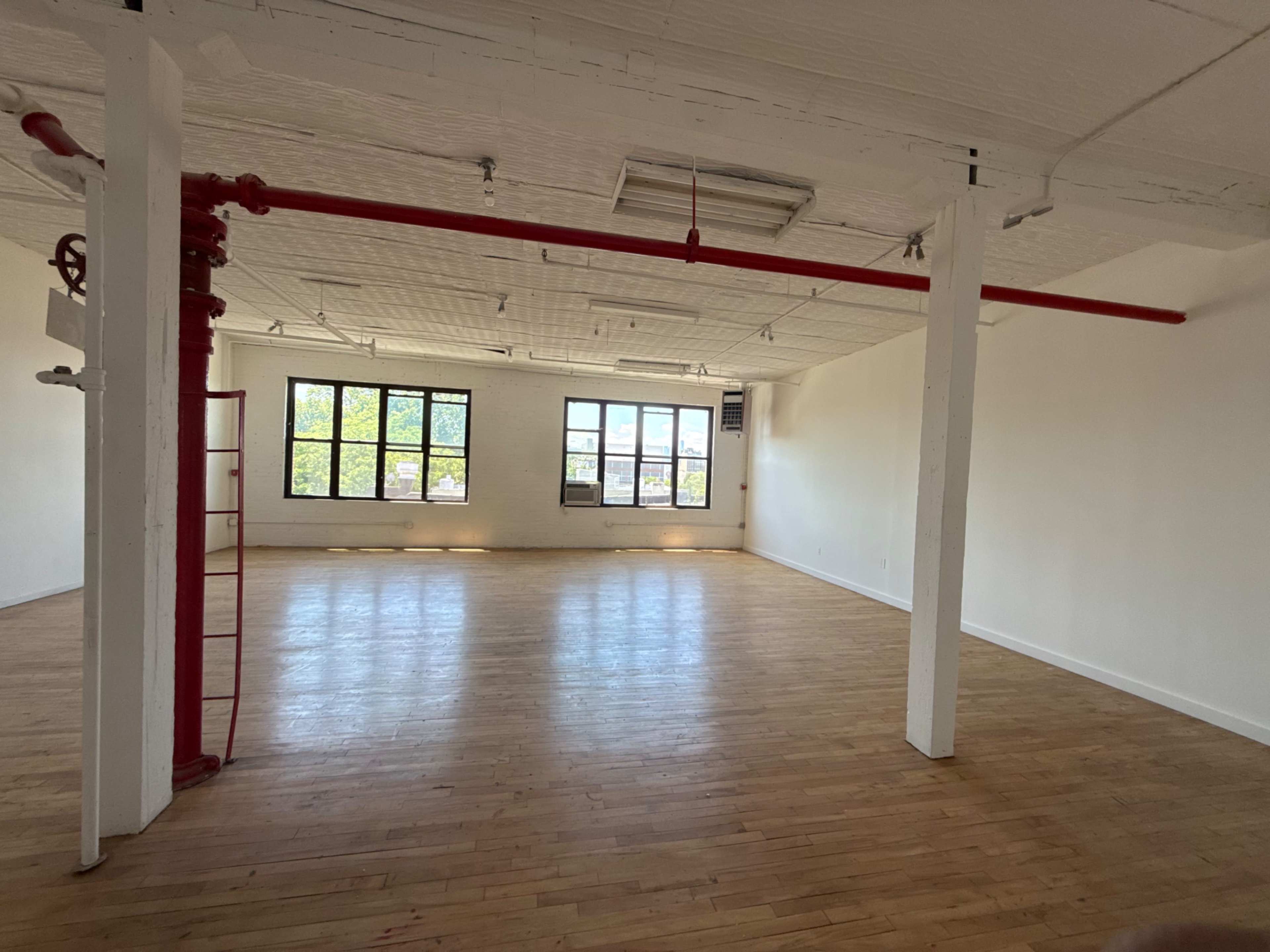 The image shows a spacious, empty room with hardwood flooring and large windows allowing natural light to enter.
